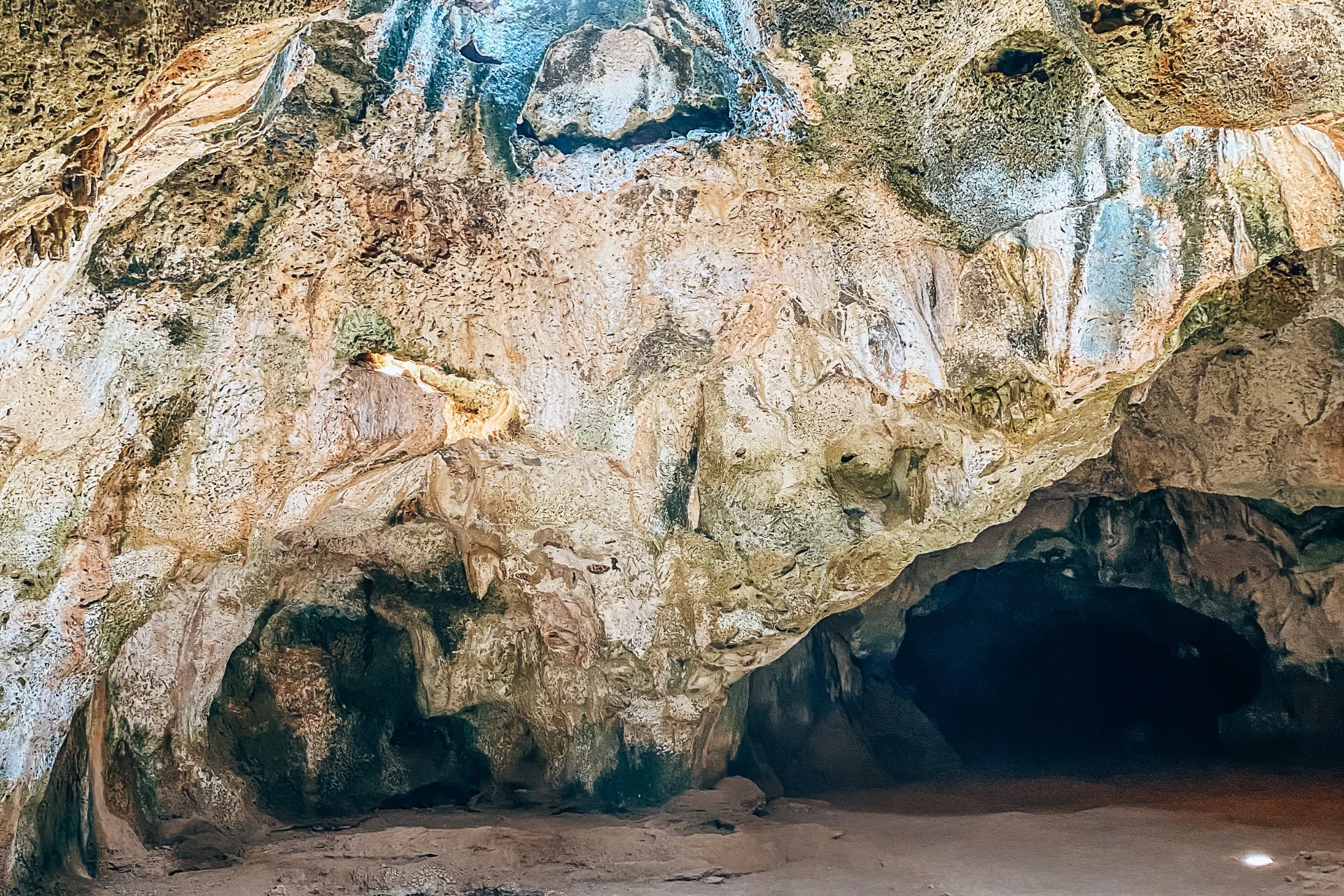 "Ingang van een natuurlijke kalksteengrot met ruwe rotsformaties in beige en groene tinten, donkere opening die leidt naar het binnenste van de grot, omgeven door verweerde texturen en lichte schaduwen, een indrukwekkende plek voor natuur- en avontuurlijke excursies.