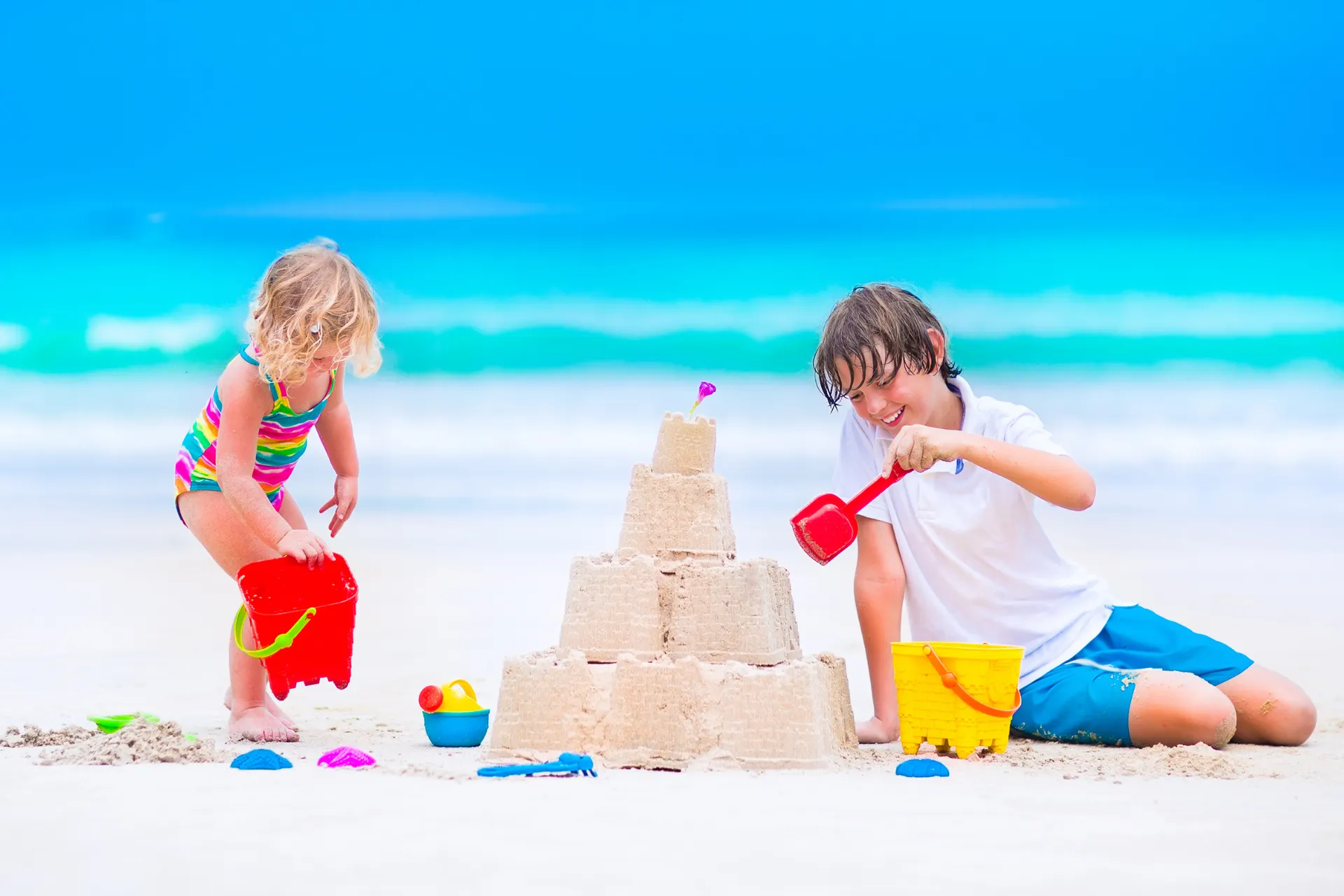 Twee kinderen bouwen een groot zandkasteel op een wit zandstrand met helder turquoise zeewater op de achtergrond. Het zandkasteel heeft meerdere lagen en torens, terwijl kleurrijke emmers en schepjes verspreid liggen rondom. De kinderen dragen zomerse kleding en genieten van een speelse strandactiviteit onder een stralend blauwe lucht. Perfect voor een familievakantie aan zee, ideaal voor ontspanning, zon en plezier tijdens een zomerse strandvakantie.