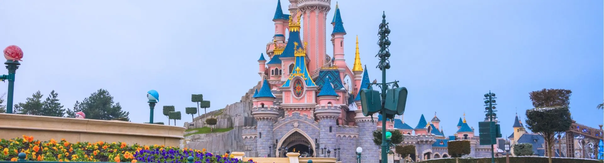 Een kleurrijke foto van Disneyland met een groot, sprookjesachtig kasteel op de achtergrond, omringd door groene tuinen en bloeiende bloemen. Het kasteel heeft hoge torens met blauwe daken en gouden accenten. Er is een heldere blauwe lucht. Het geheel straalt vreugde uit.