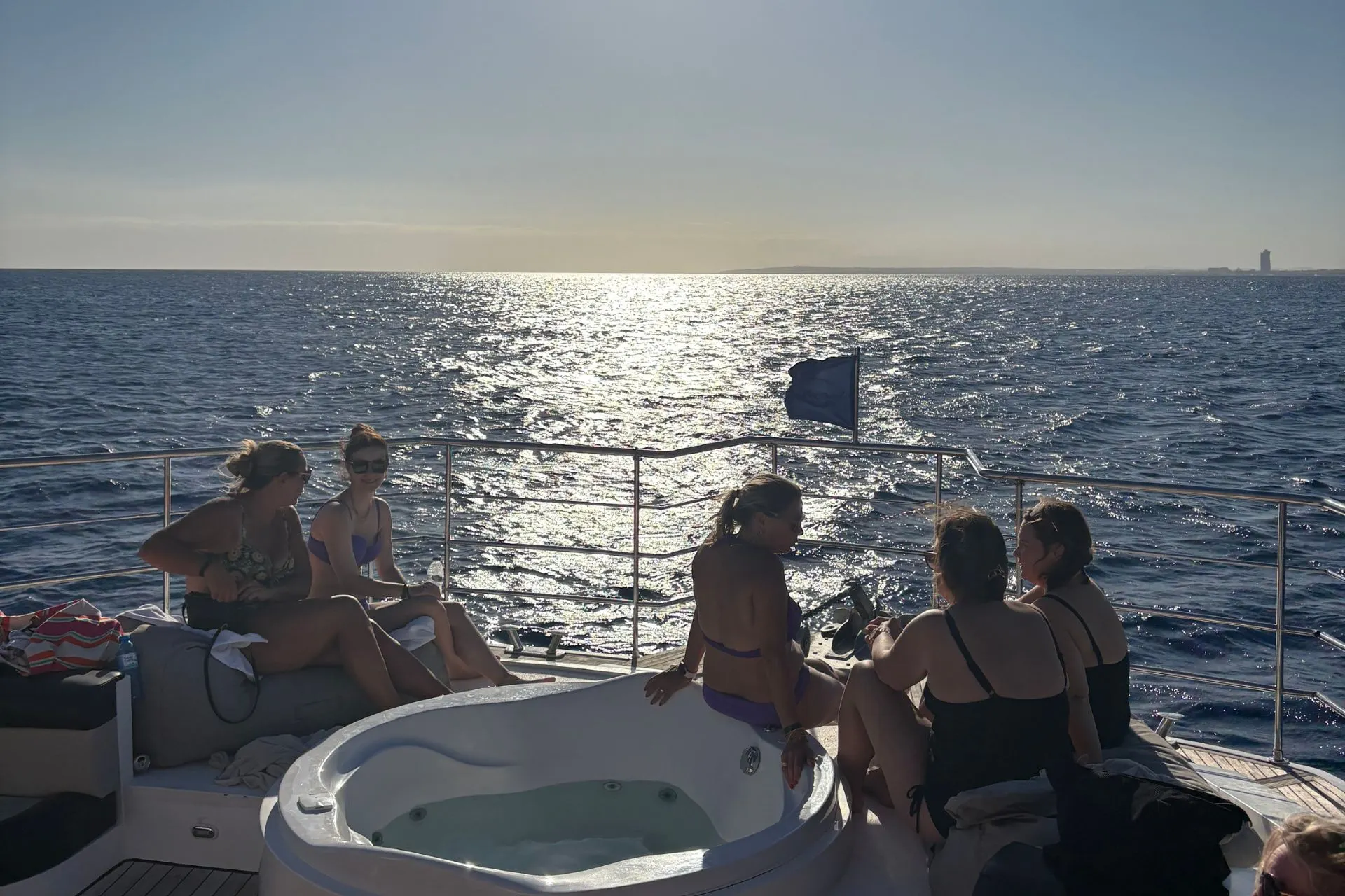 Vier personen zitten in badkleding bij een jacuzzi op het dek van een boot, met uitzicht op de zon die schittert op de zee.