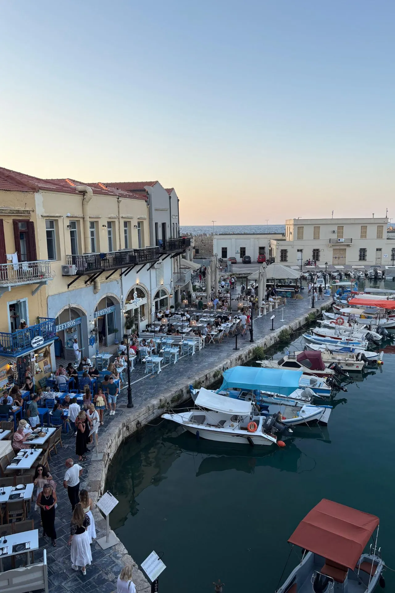 Gezellige haven van Rethymnon met terrasjes langs het water, dobberende bootjes en warme avondzon boven de historische gebouwen.