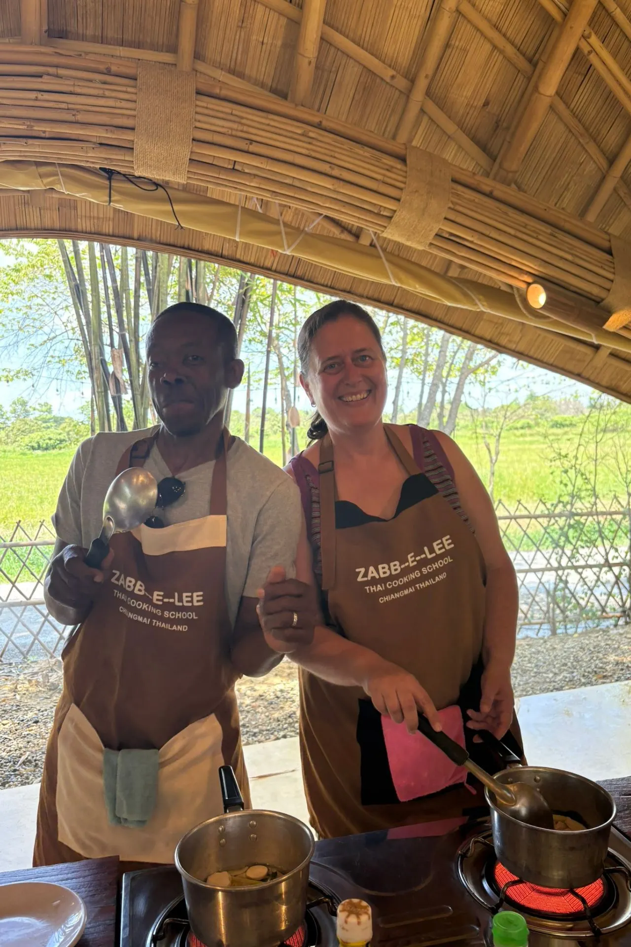 Twee mensen dragen schorten van Zabb-E-Lee Thai Cooking School en bereiden gerechten op een kookstation in een open bamboepaviljoen.