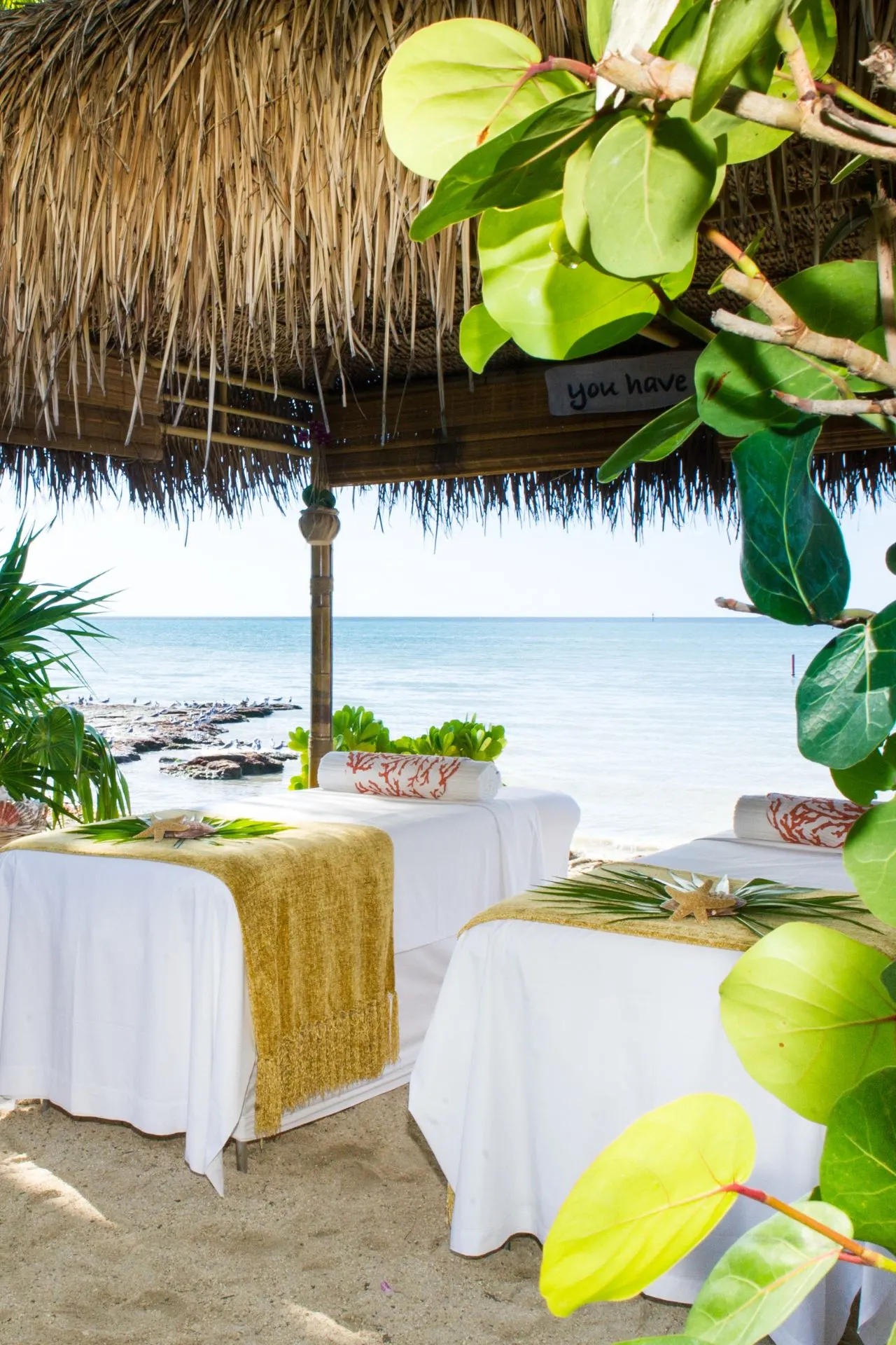 Buitenspa op het strand met rieten dak, comfortabele massagetafels en uitzicht op een kalme zee.