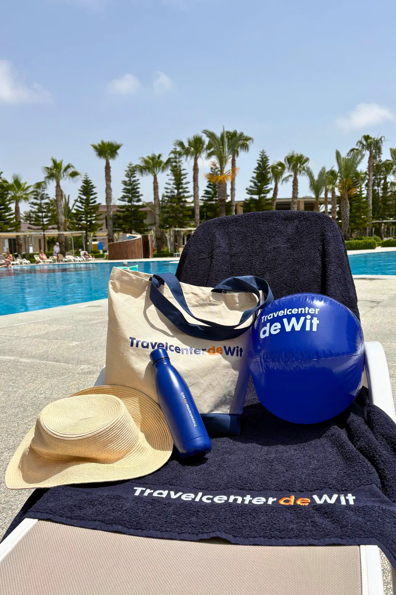 Ligbed bij het zwembad van Sunmelia Beach Resort in Turkije met Travelcenter de Wit-strandartikelen, waaronder een strandtas, handdoek, waterfles en strandbal. Op de achtergrond staan palmbomen, ligbedden en het grote zwembad van het resort aan de Turkse Rivièra, ideaal voor een ontspannen all inclusive vakantie.
