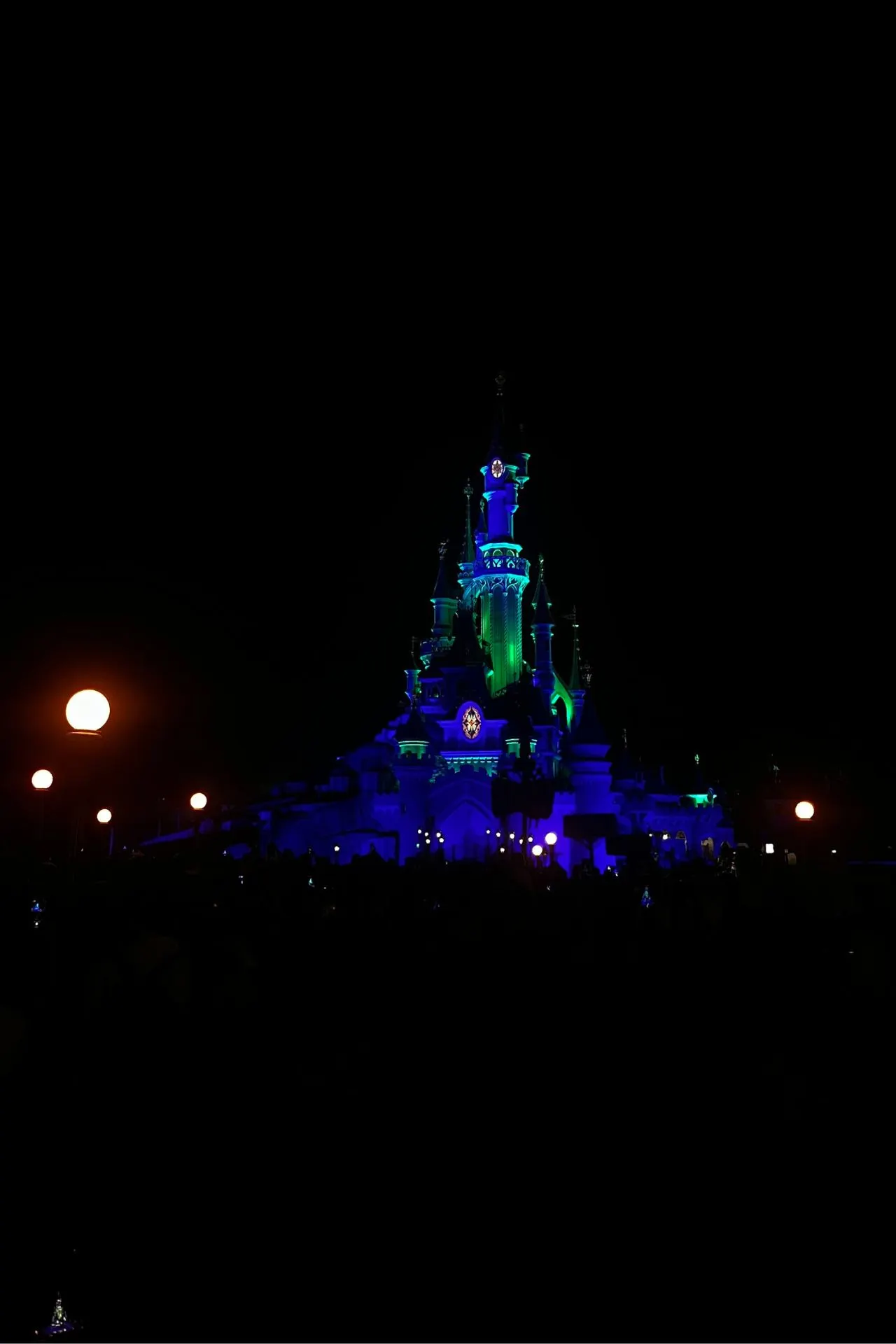 Doornroosje kasteel in Disneyland Parijs verlicht in groene en paarse tinten tijdens de avondshow, omringd door donkere nacht en warme lichtbollen.