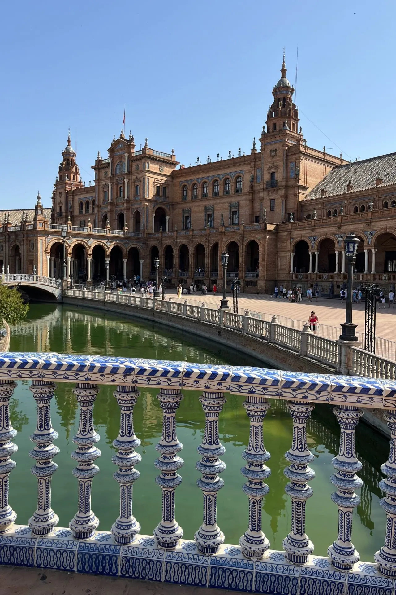 Uitzicht op het centrale gebouw van Plaza de España in Sevilla, Spanje, met zijn halfronde structuur, torens en booggalerijen. Op de voorgrond een typisch Spaans betegeld bruggetje over het kanaal met groen water.