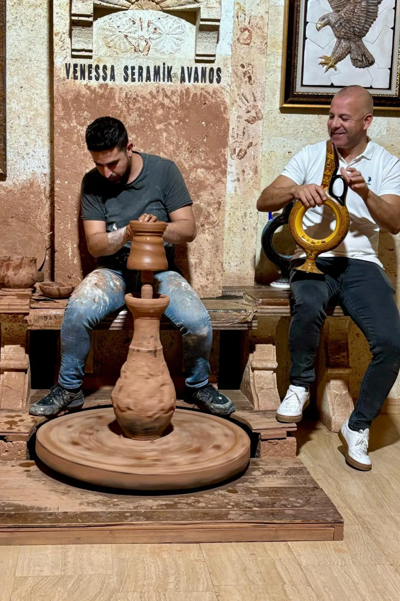 Twee mannen demonstreren traditioneel pottenbakken in een werkplaats in Avanos, Cappadocië, Turkije. Op de voorgrond draait een grote kleischijf waarop een vaas wordt gevormd, terwijl de pottenbakker geconcentreerd werkt met zijn handen. Naast hem bespeelt een muzikant een traditioneel instrument, wat zorgt voor een authentieke sfeer. De achtergrond toont aardetinten, keramische kunstwerken en decoratieve elementen die de rijke ambachtelijke cultuur van deze regio benadrukken. Populaire activiteit voor toeristen en liefhebbers van handwerk.