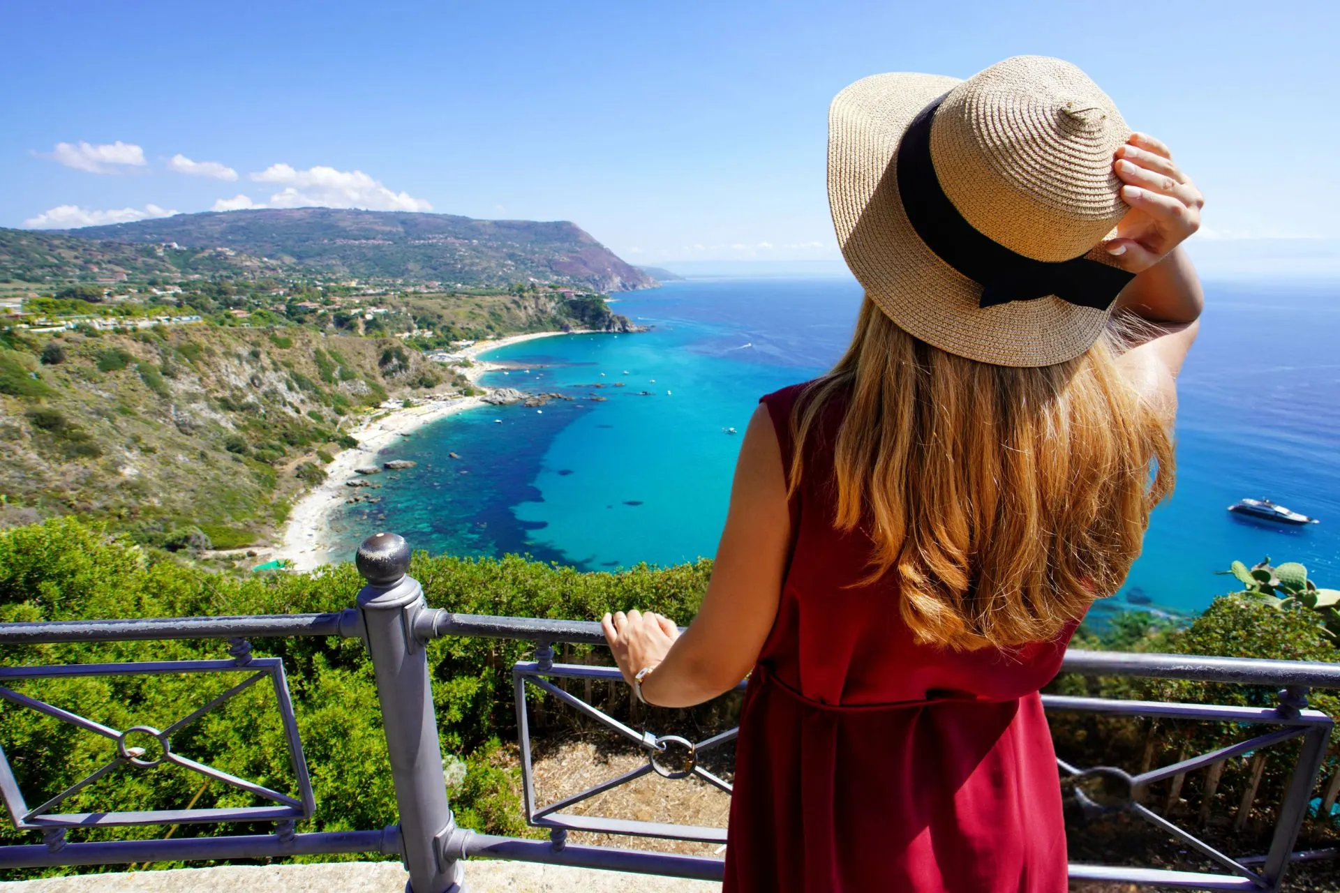 Vrouw met een zonnehoed kijkt uit over een helderblauwe kustlijn en groene heuvels vanaf een uitzichtpunt.
