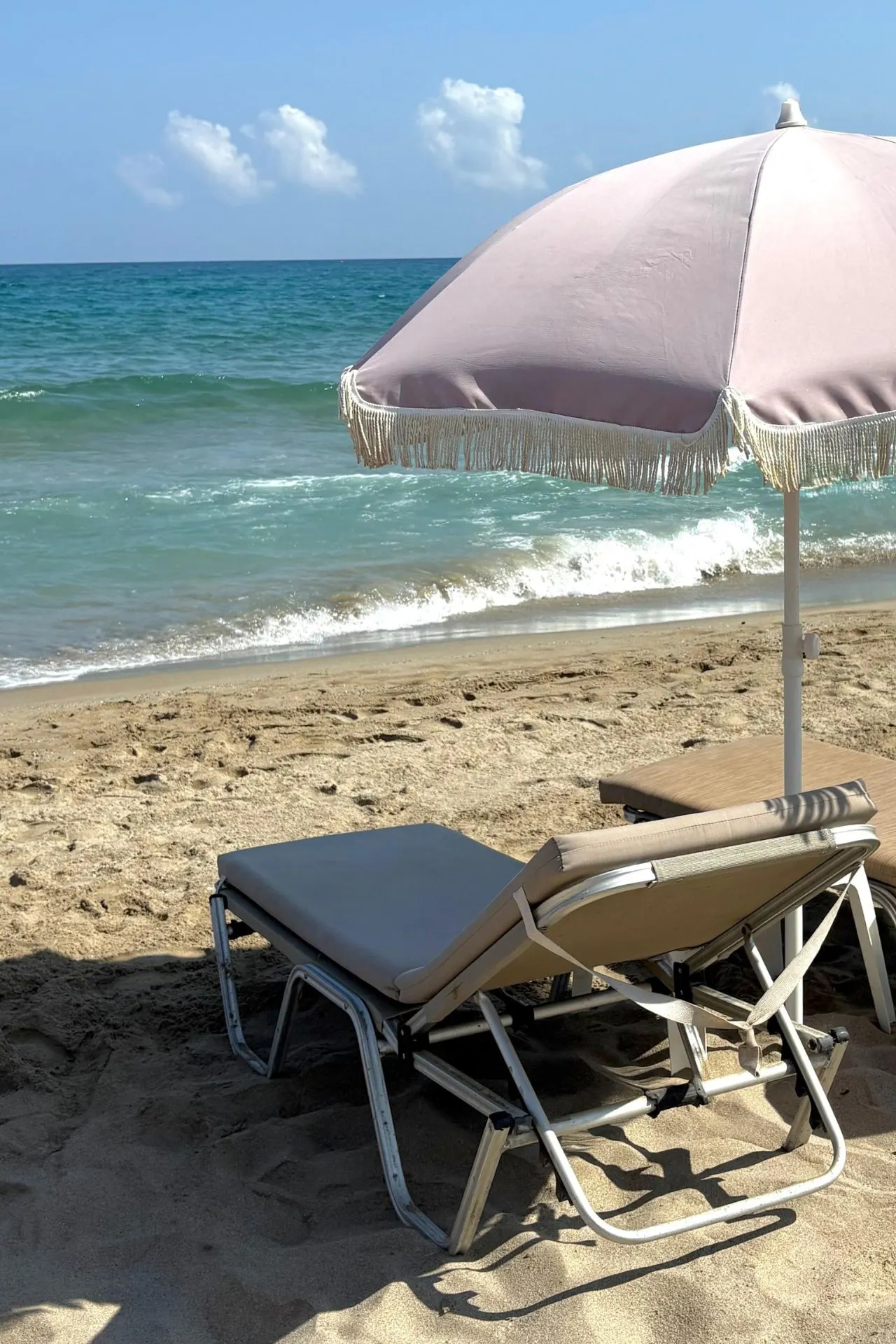 Twee ligbedden met parasol op het zandstrand voor het Batis Beach Hotel, met helderblauw zeewater en rustige golven op de achtergrond.
