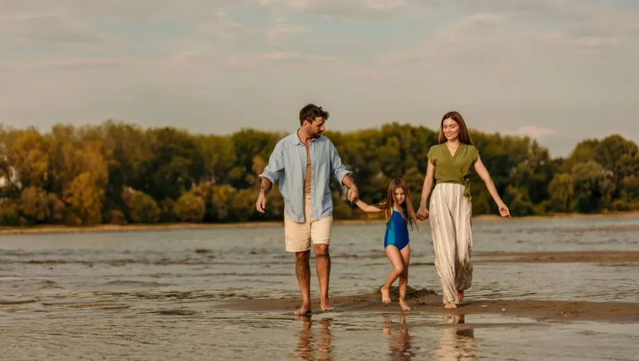 Een jong gezin van drie loopt hand in hand door ondiep water tijdens een ontspannen wandeling langs een rivier of meer. De vader draagt een lichtblauw overhemd en korte broek, de moeder draagt een groene top en een losse, gestreepte zomerse broek, en het meisje in het midden draagt een blauwe badpak. Ze kijken elkaar liefdevol aan terwijl ze door het kalme water stappen, met op de achtergrond een bosrijke oever vol groene bomen en een heldere lucht in zachte avondkleuren. Deze sfeervolle vakantiefoto ademt rust, verbondenheid en natuurbeleving – ideaal voor marketing rond gezinsvriendelijke vakanties, natuurcampings, slow travel of vakantie boeken in landelijke bestemmingen. Zoekwoorden: gezinsvakantie in de natuur, rustgevende reizen, wandelen tijdens vakantie, kindvriendelijke vakantiebestemming boeken.