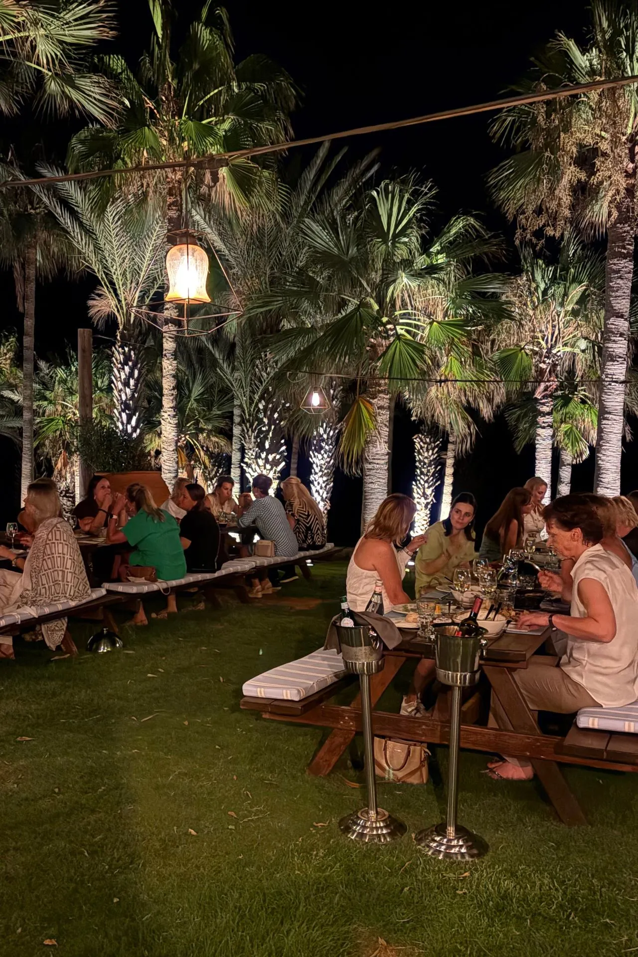 Buitenrestaurant in een tuin met palmbomen, waar meerdere tafels zijn gedekt voor een avondmaal, sfeerverlichting en mensen die dineren.