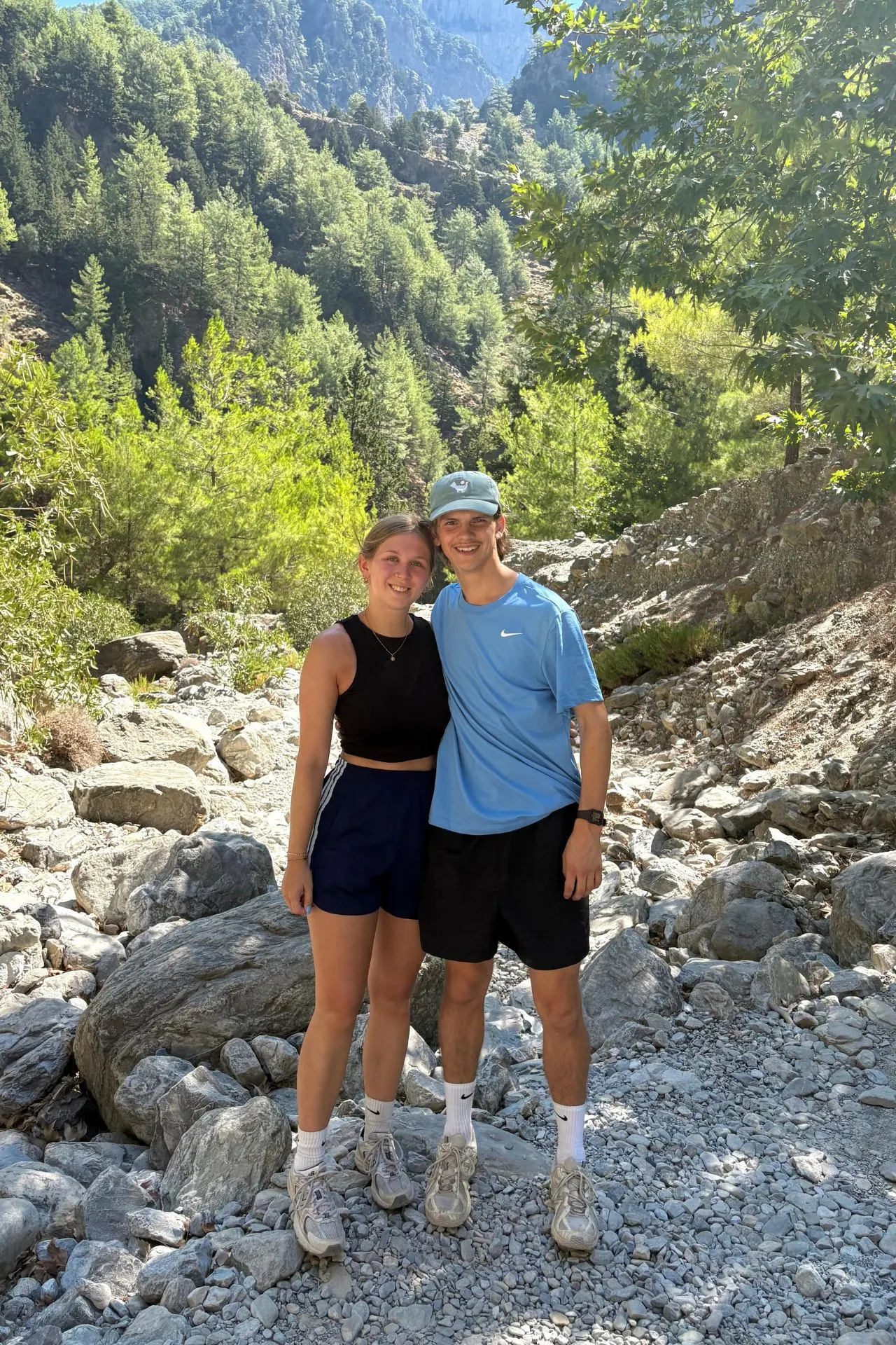 Twee wandelaars poseren lachend tussen de rotsen en dennenbomen in de Samariakloof, met zonlicht dat door de bergen heen valt.