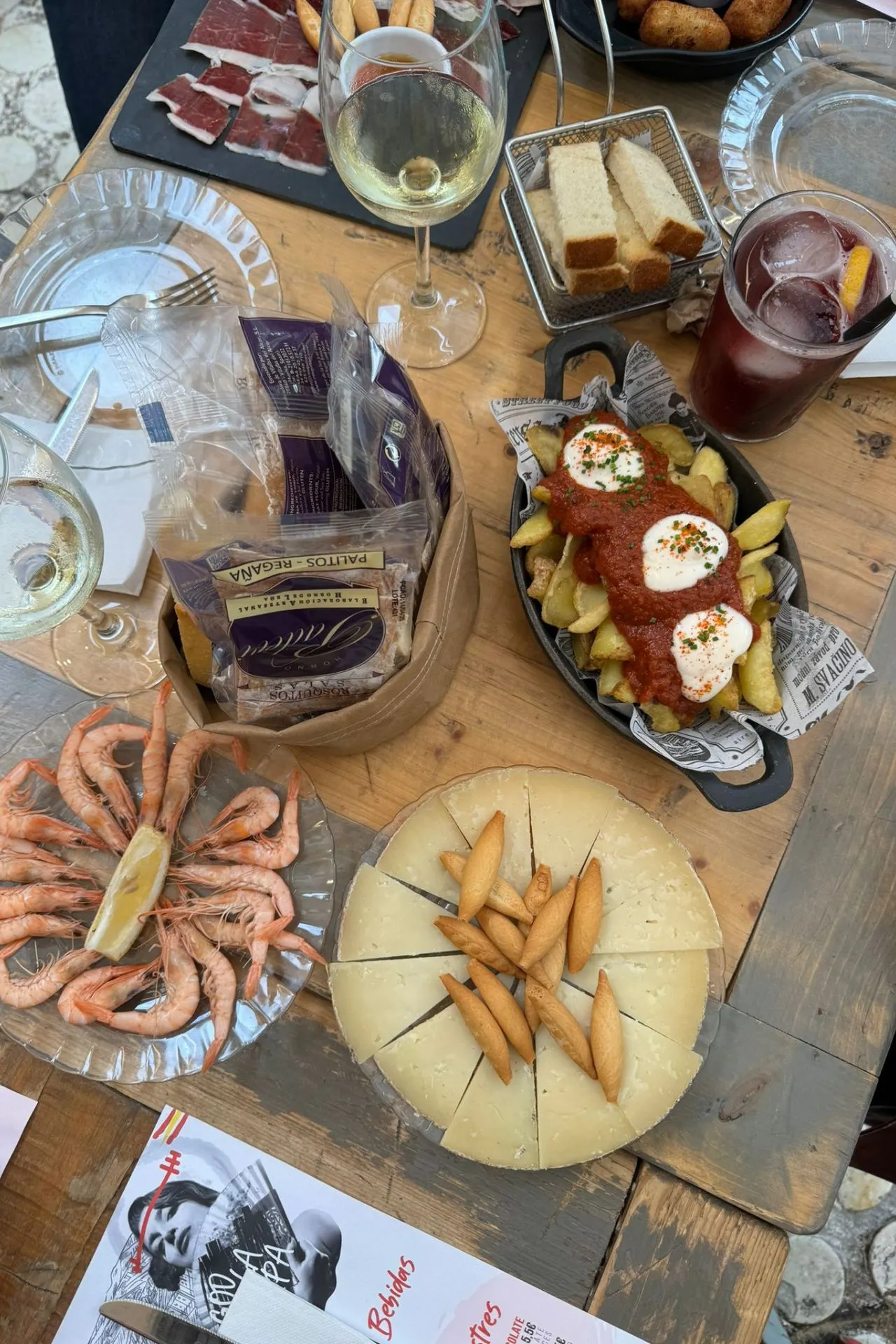 Close-up van een tafel vol met Spaanse tapas, inclusief garnalen, kaas, patatas bravas, en diverse drankjes zoals sangria en witte wijn.