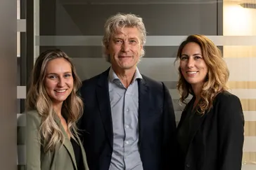 Emmie, Ad en Michelle de Wit staan naast elkaar, lachen en stralen samen in het reisbureau van Travelcenter de Wit, waarbij hun positieve energie en samenwerking duidelijk zichtbaar zijn.