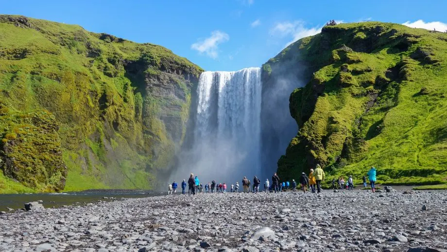 Toeristen wandelen naar de voet van de Skógafoss-waterval in IJsland, waar het water krachtig naar beneden stort tegen een met mos begroeide klif.