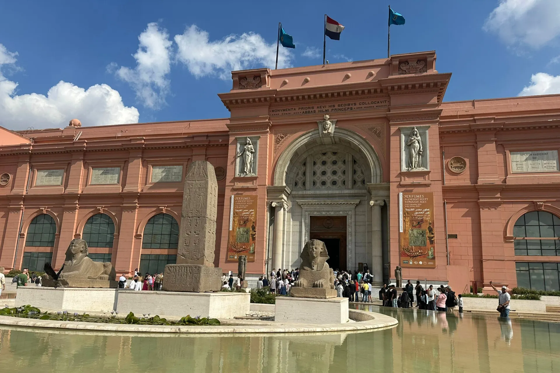 Buitenaanzicht van het Egyptisch Museum in Caïro met bruinrode stenen gevel, drie wapperende vlaggen op het dak en een klein waterpartij op de voorgrond.