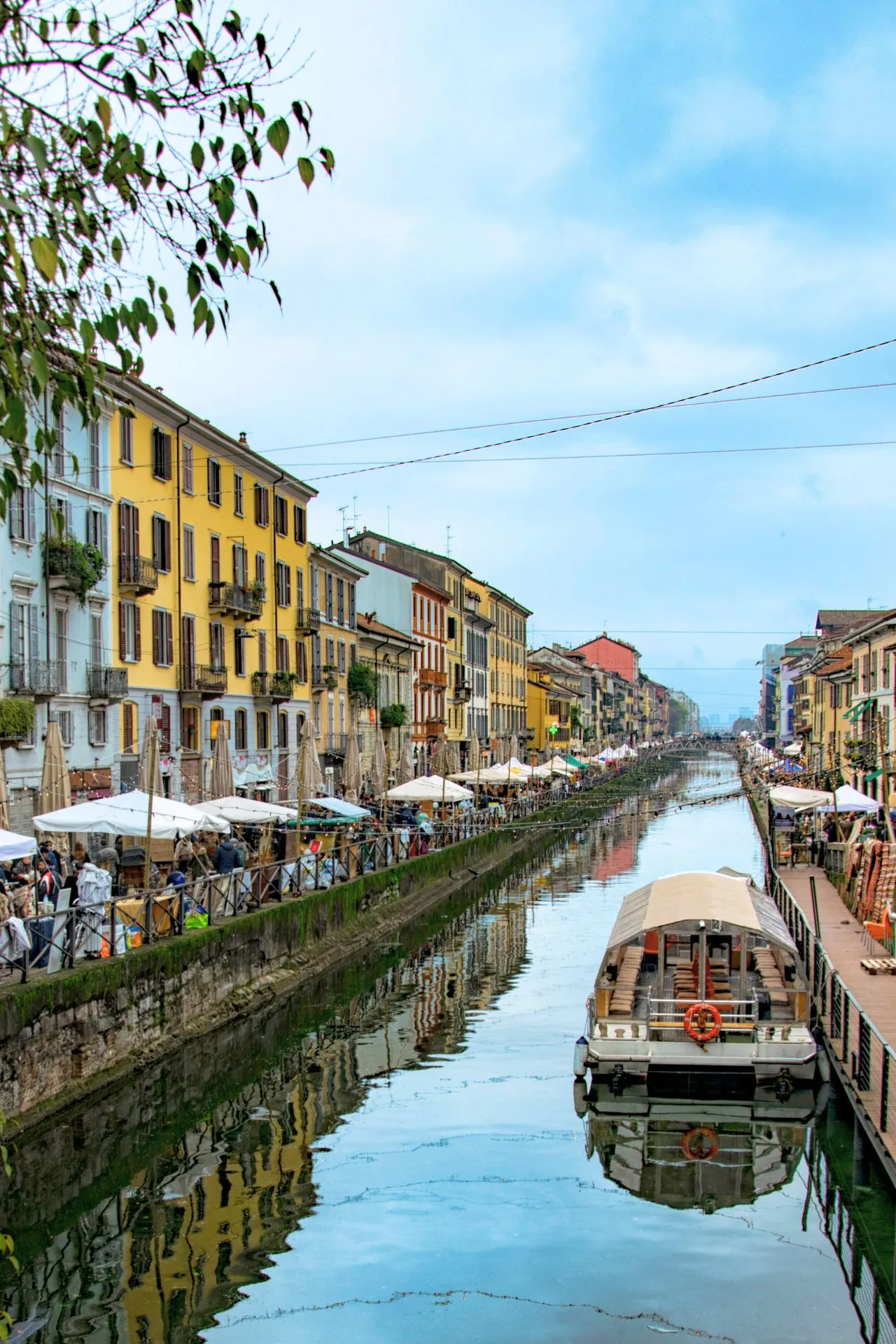 Zicht op het Navigli-kanaal in Milaan, met kleurrijke gebouwen aan weerszijden, een boot op het water en marktkraampjes en terrasjes langs de promenade.
