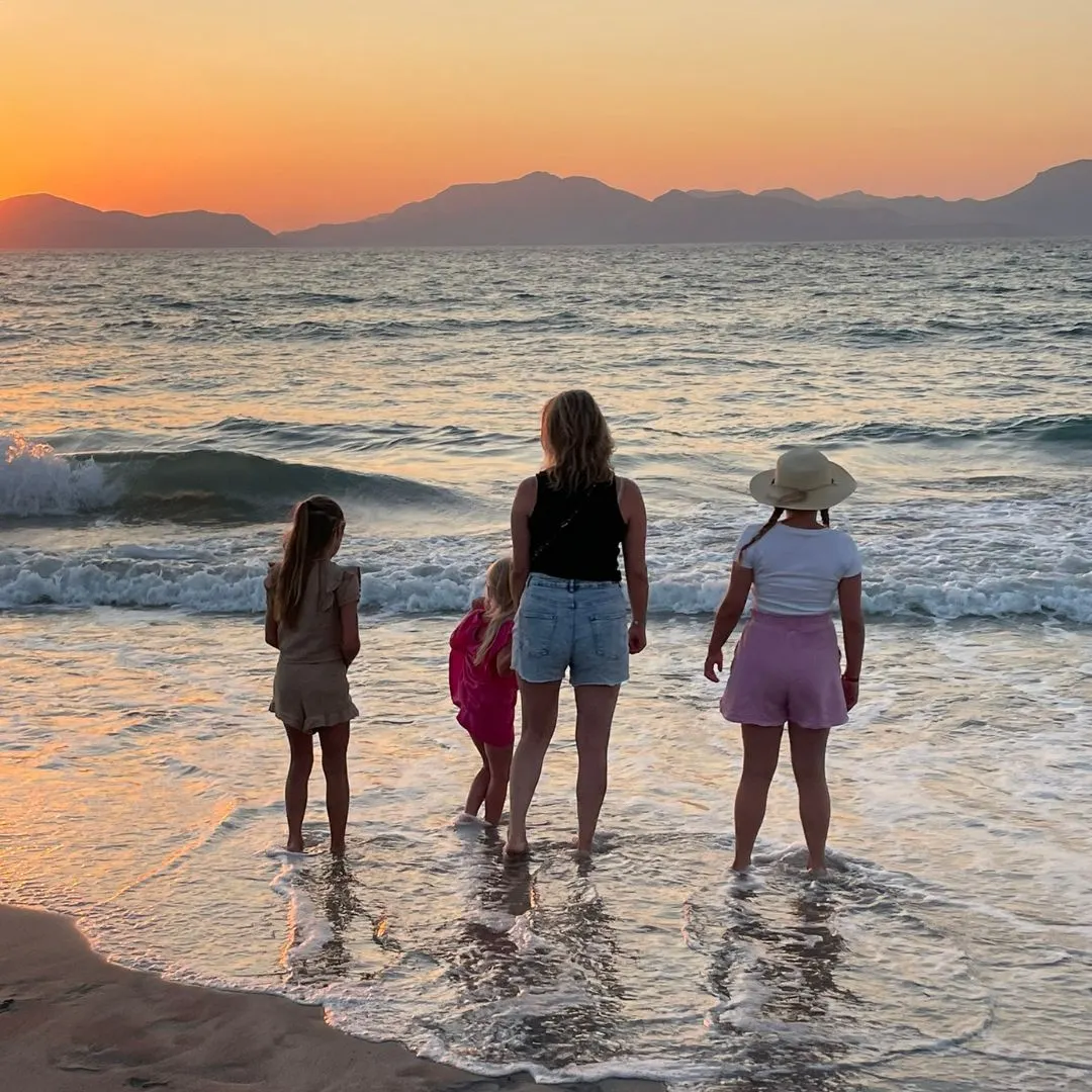 Moeder en drie kinderen staan met de voeten in het water op het strand van Kos, terwijl de zon langzaam achter de bergen verdwijnt