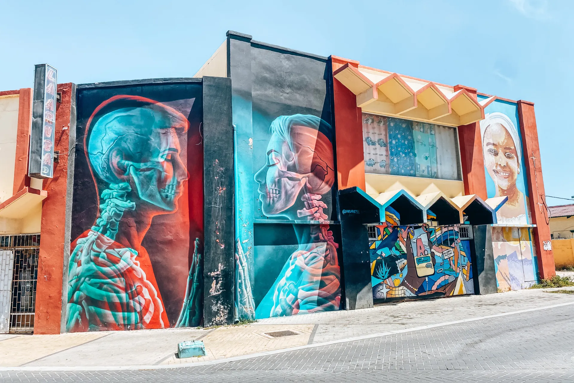 "Levendige muurschildering op een gebouw met anatomische illustraties van menselijke skeletten en organen in rood en blauw tinten, gecombineerd met abstracte kunst en geometrische vormen, gelegen langs een straat onder een heldere blauwe lucht."