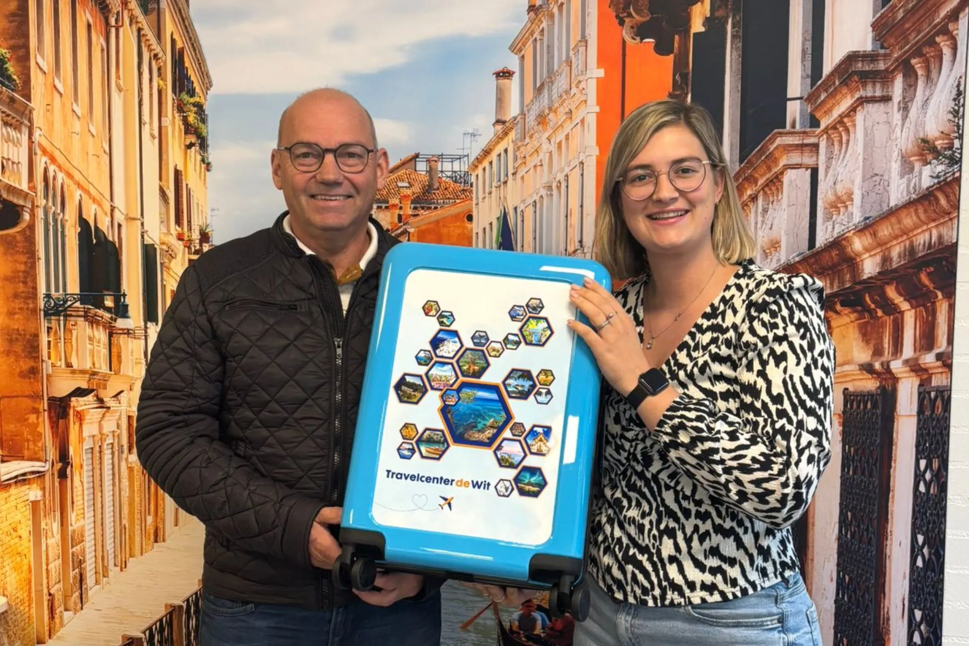 Man en vrouw houden samen een blauwe koffer vast met Travelcenter de Wit-logo tijdens de prijsuitreiking van de winactie bij Travelcenter de Wit Gemert.