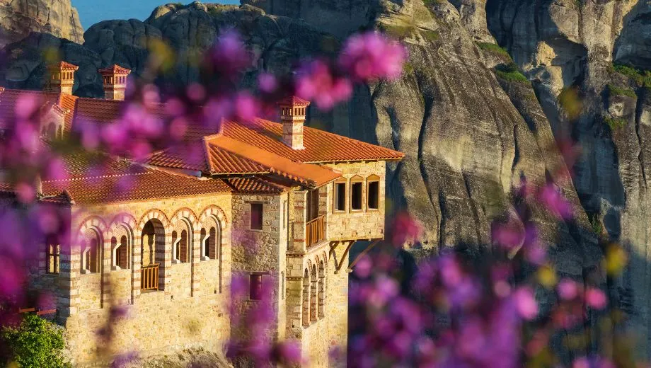 "Close-up van een Meteora-klooster in Griekenland, gebouwd op een hoge rotsformatie, omringd door paarse bloemen op de voorgrond. Het klooster heeft een traditioneel rood dak en stenen muren, typerend voor de orthodoxe architectuur. Meteora is een UNESCO-werelderfgoed en een populaire bestemming voor cultuur, natuur en spiritualiteit. Ontdek indrukwekkende kloosters, adembenemende uitzichten en wandelroutes in Centraal-Griekenland."