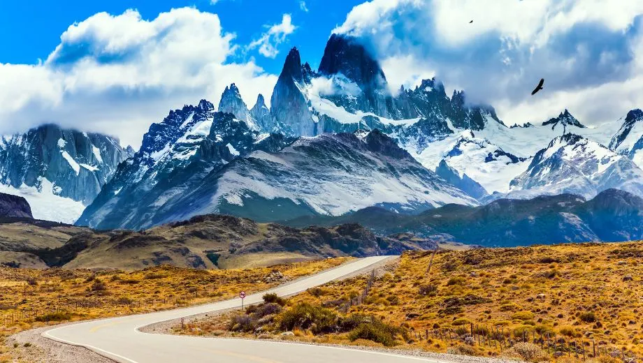 Panoramisch uitzicht over Patagonië in Argentinië met een kronkelende weg door een uitgestrekt landschap. Op de achtergrond rijzen indrukwekkende besneeuwde bergtoppen van de Andes op onder een heldere blauwe lucht met wolken. Een iconisch decor voor een rondreis door Chili en Argentinië.