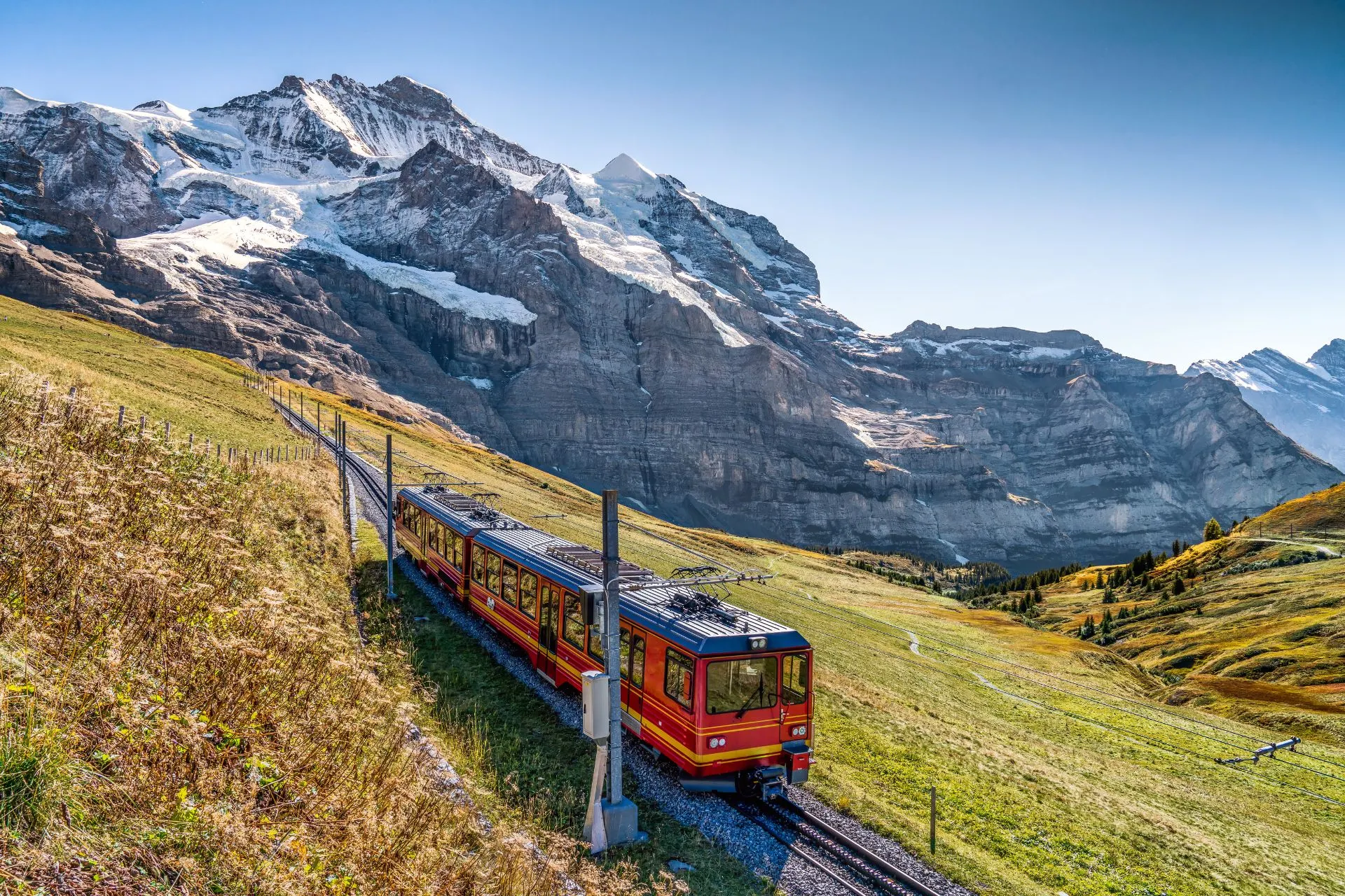 Rood-gele trein rijdt door de Zwitserse Alpen met uitzicht op besneeuwde bergtoppen en groene valleien.