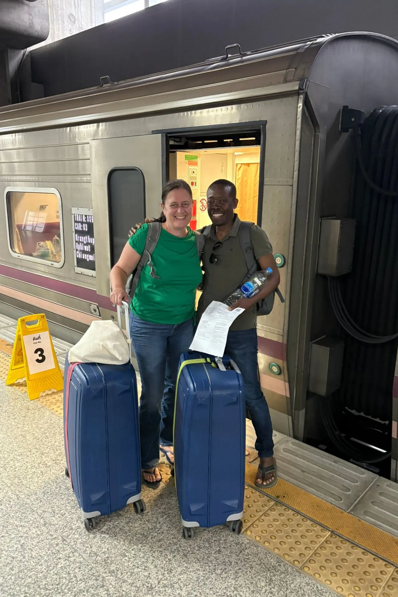 Twee reizigers glimlachen bij de ingang van een nachttrein van Bangkok naar Chiang Mai, elk met een blauwe koffer en rugzak.