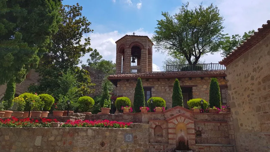"Traditionele binnenplaats van een Meteora-klooster in Griekenland, met een stenen klokkentoren, verzorgde tuin en bloeiende planten in terracotta potten. Deze historische plek weerspiegelt de orthodoxe cultuur en architectuur van Meteora, een UNESCO-werelderfgoed. Perfect voor een culturele reis naar Griekenland, ontdek kloosters, spirituele tradities en prachtige natuur in een serene omgeving."