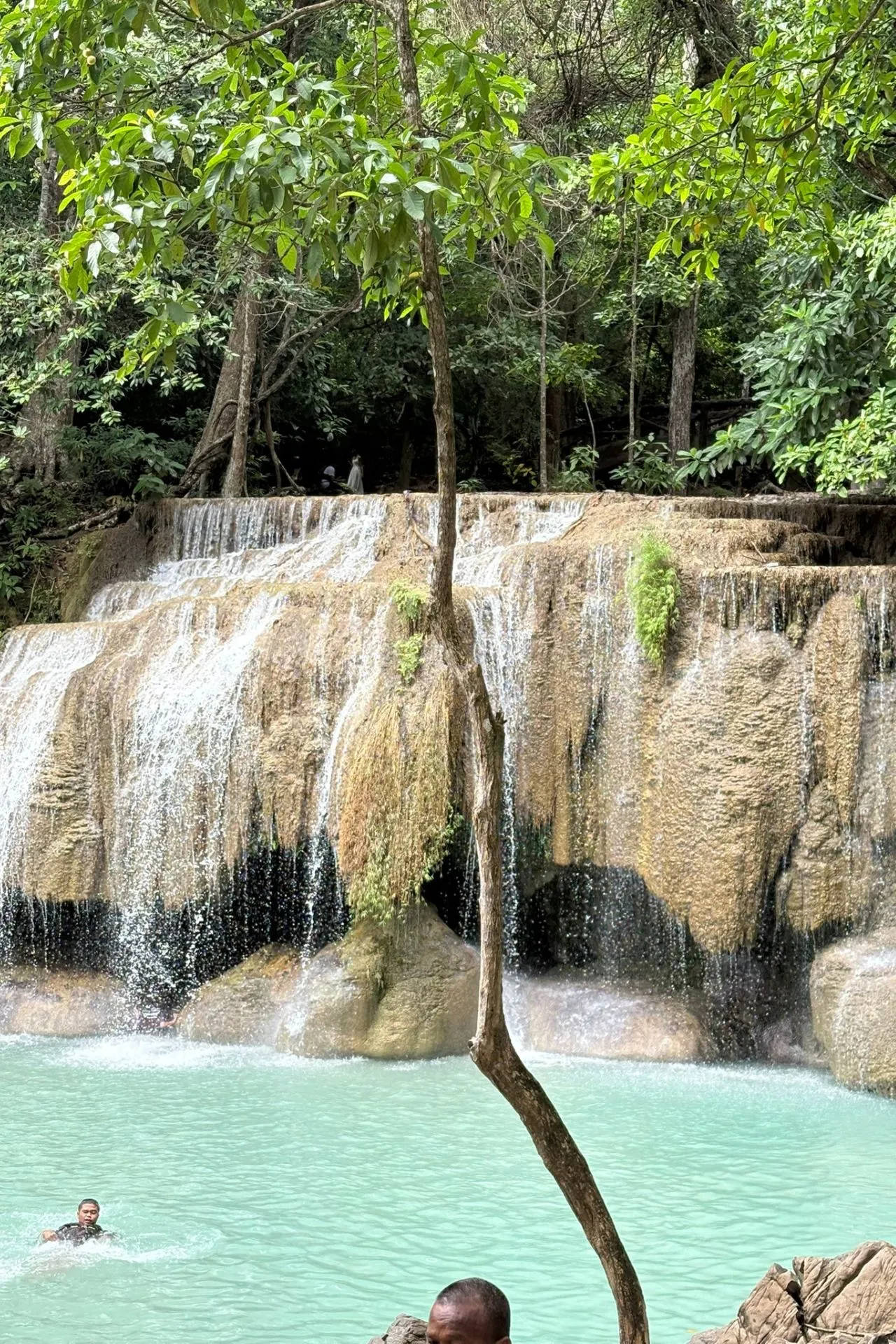 Een brede, trapsgewijze waterval met helder turquoise water, omgeven door weelderig groen, met mensen die in het water zwemmen.