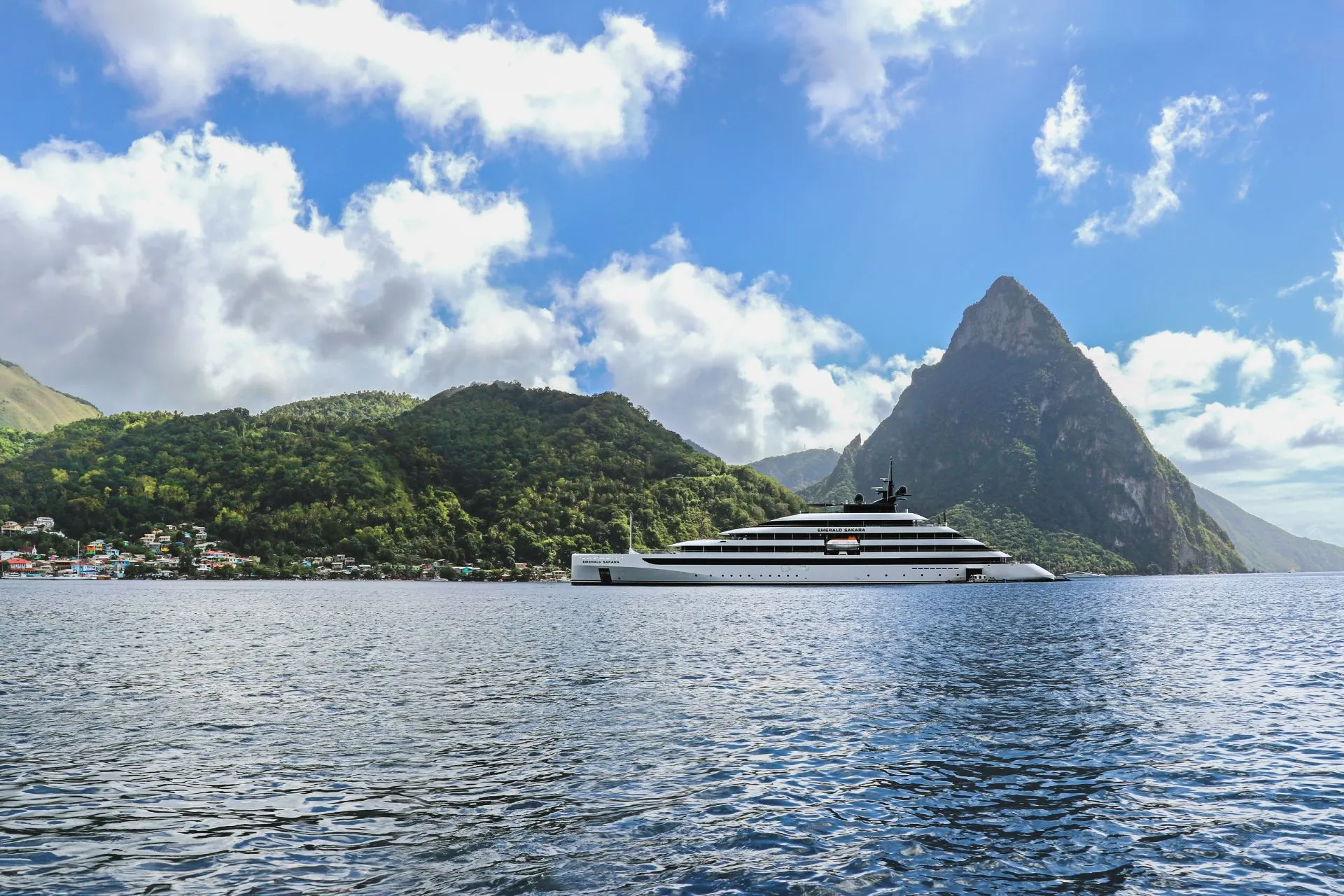 Een indrukwekkend modern jacht vaart voor een spectaculair tropisch landschap met groene heuvels en steile bergtoppen, omringd door helderblauw water. De serene omgeving en het elegante design van het schip benadrukken de exclusieve sfeer van een luxe zeecruise. Deze idyllische bestemming onderstreept de unieke reizen die luxe cruise maatschappijen bieden, gericht op comfort, ontdekkingen en uitzonderlijke natuurervaringen.

