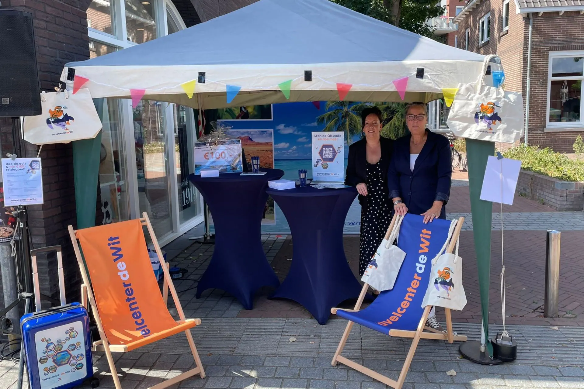 Promotiekraam van Travelcenter de Wit tijdens Thuis in Rosmalen 2025, met twee medewerkers achter een stand met banners, stoelen met logo en goodiebags.