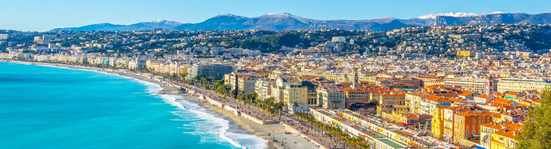 Panoramisch uitzicht op een kuststad met een lange boulevard langs een helderblauwe zee. Aan de kustlijn staan rijen gebouwen met rode daken en lichte gevels, afgewisseld met palmbomen die een mediterrane sfeer creëren. Op de achtergrond liggen groene heuvels en bergen met besneeuwde toppen, wat een prachtig contrast vormt met de zonnige kust. Deze foto benadrukt een populaire vakantiebestemming, ideaal voor strandwandelingen, cultuur en ontspanning in een schilderachtige omgeving.