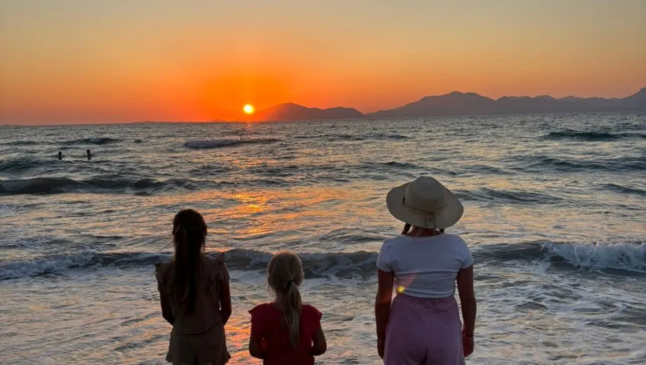 Drie meisjes staan op het strand van Kos en kijken naar de ondergaande zon boven de zee, met een warme oranje gloed op de golven