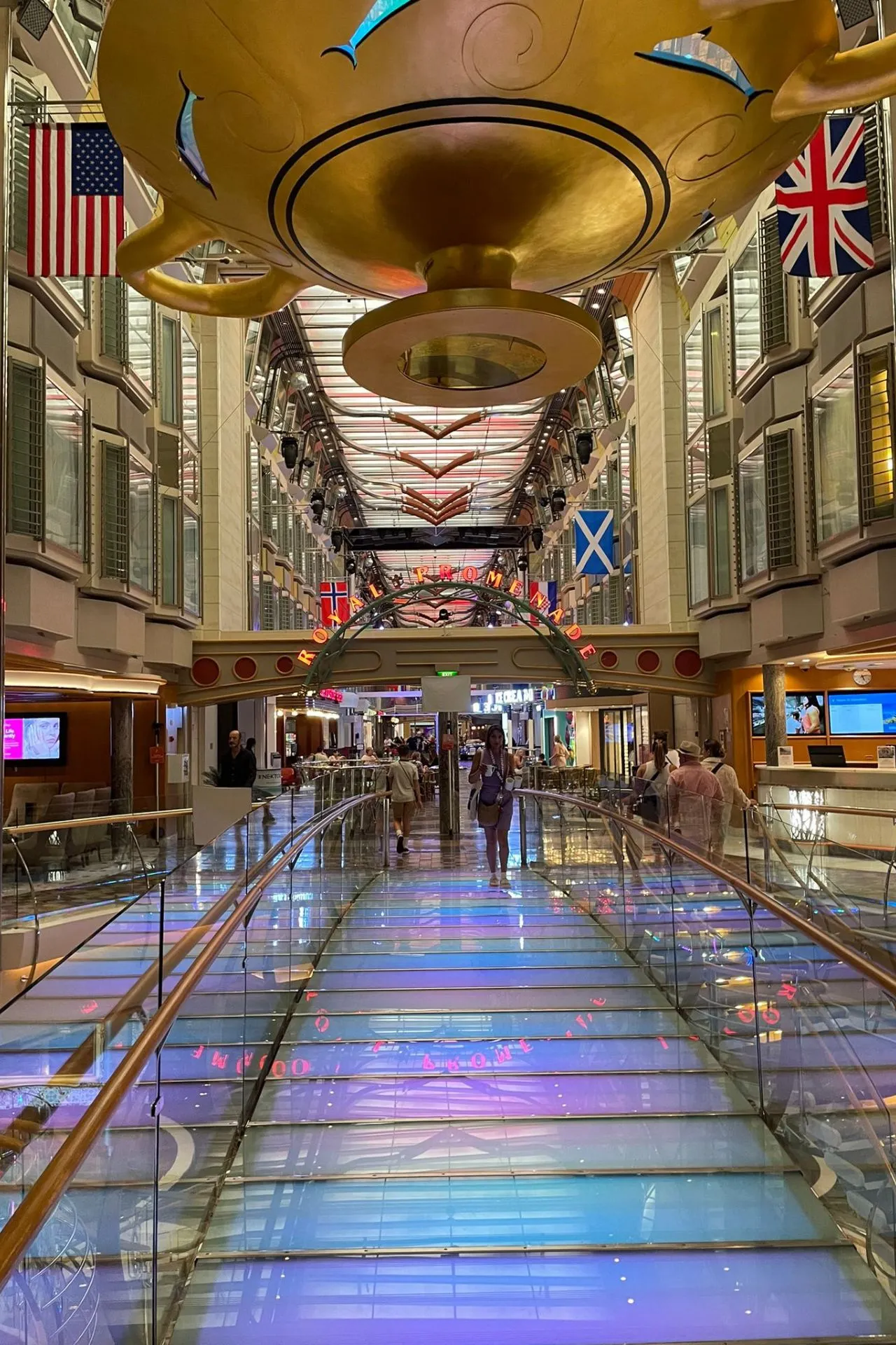 Royal Promenade aan boord van de Independence of the Seas, met glazen loopbrug, kleurrijke verlichting, vlaggen en winkels aan weerszijden.
