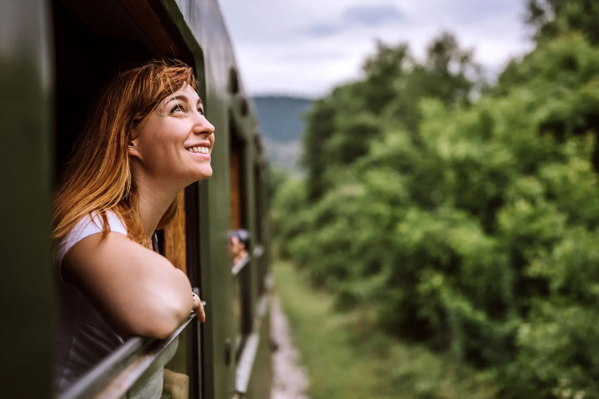 Lachende vrouw leunt uit het open raam van een groene trein en kijkt uit over het omliggende bosrijke landschap.