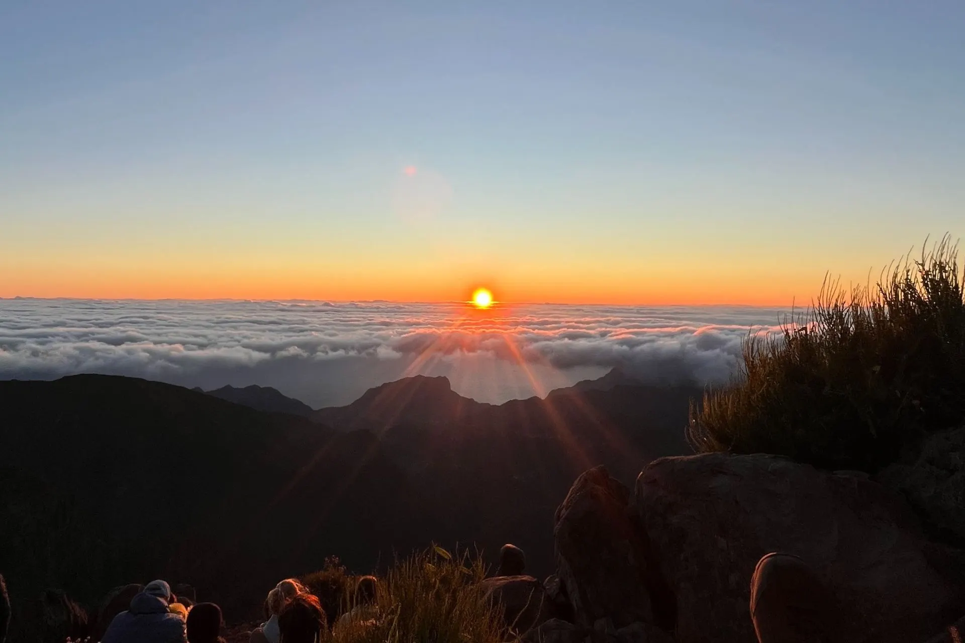 Opkomende zon boven een zee van wolken bij Pico do Arieiro. De zon straalt fel oranje licht, omringd door een blauwe lucht. In de voorgrond zitten mensen op rotsen en gras om het uitzicht te bewonderen.
