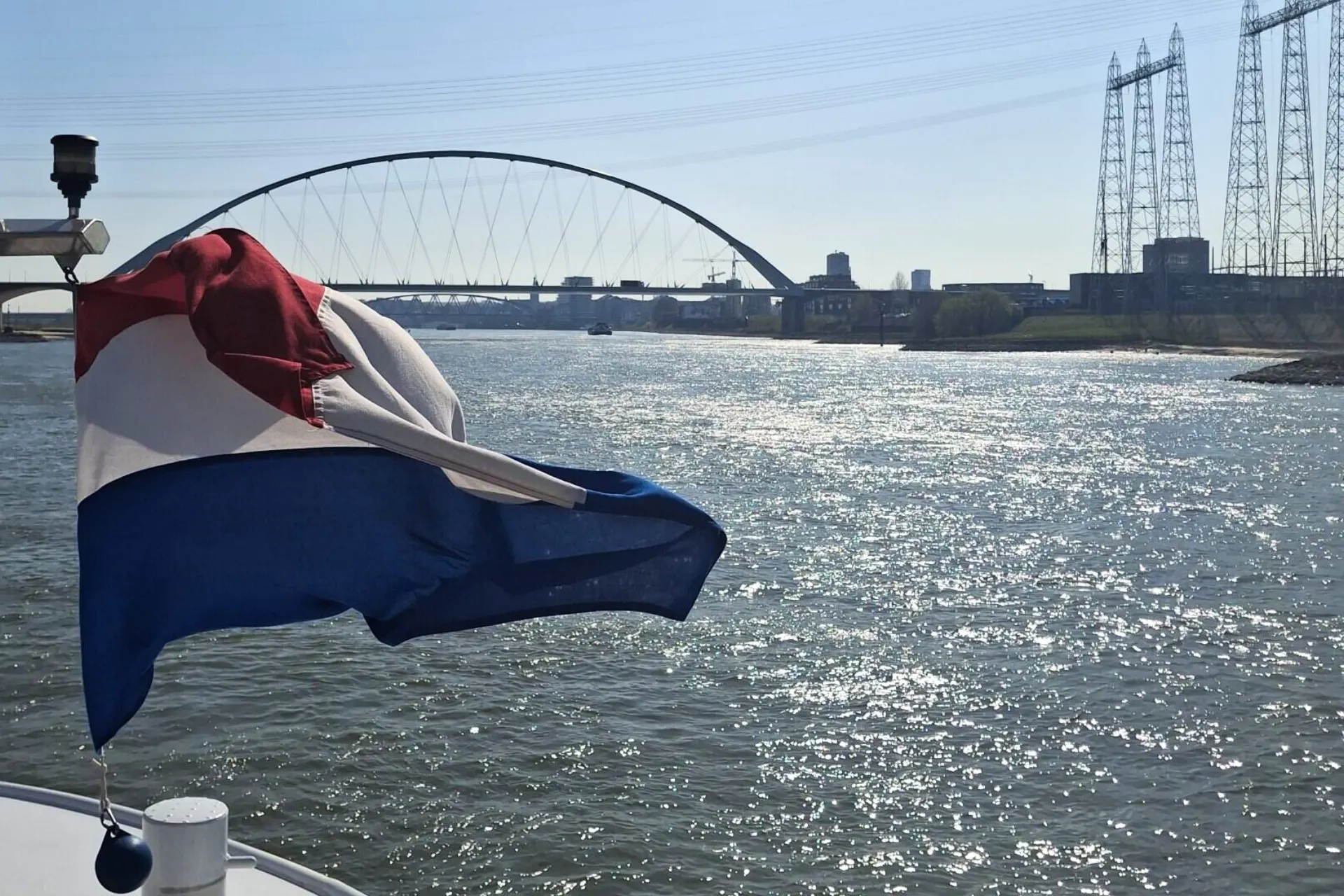 Wapperende Nederlandse vlag op het dek van de Emerald Destiny met uitzicht op de brug van Nijmegen en de rivier.