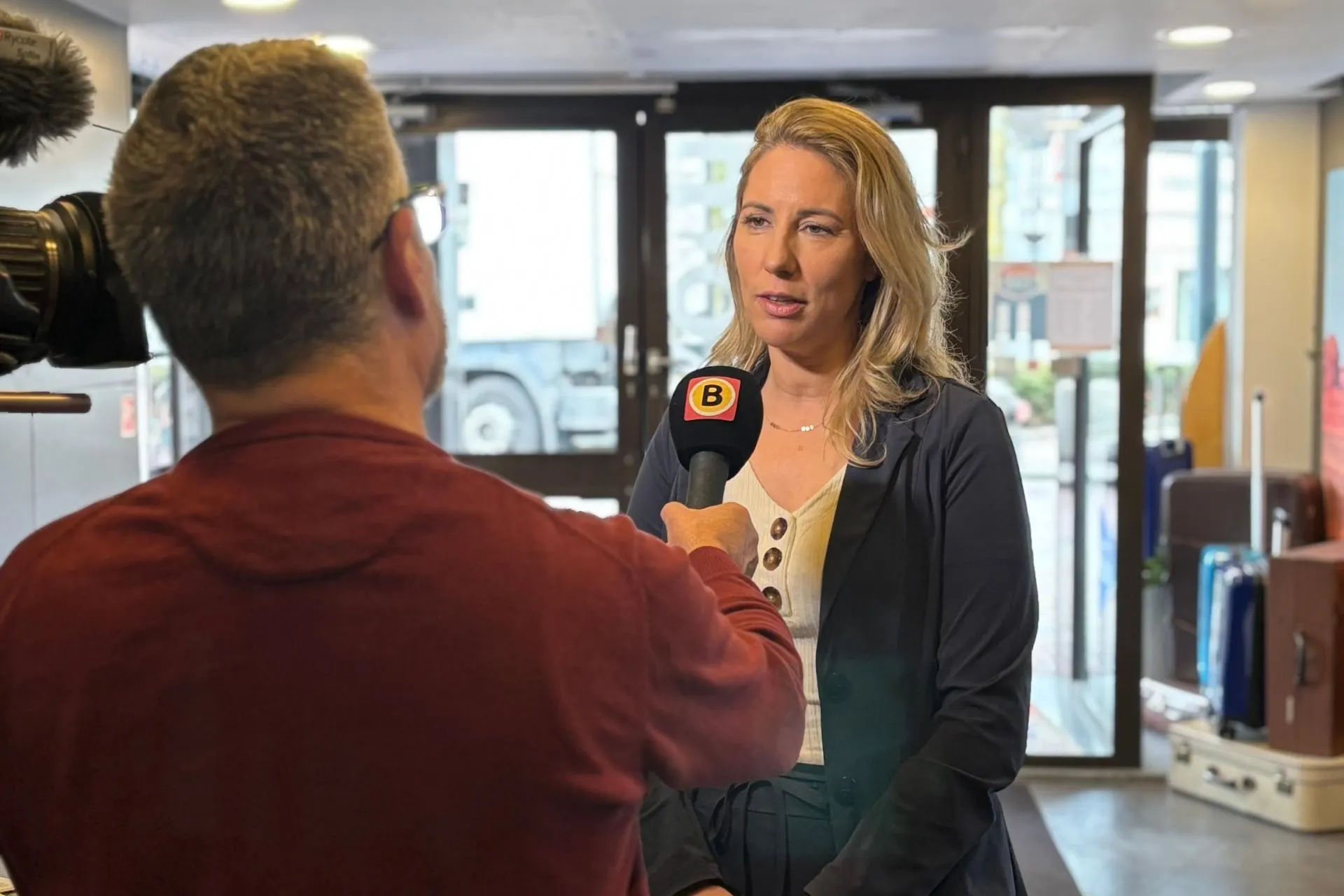  Een journalist van Omroep Brabant interviewt Michelle de Wit, eigenaresse van Travelcenter de Wit, bij de ingang van het reisbureau. De journalist houdt een microfoon met het logo van Omroep Brabant vast, terwijl een cameraman het interview filmt. Op de achtergrond zijn koffers en de glazen entree van het kantoor zichtbaar.