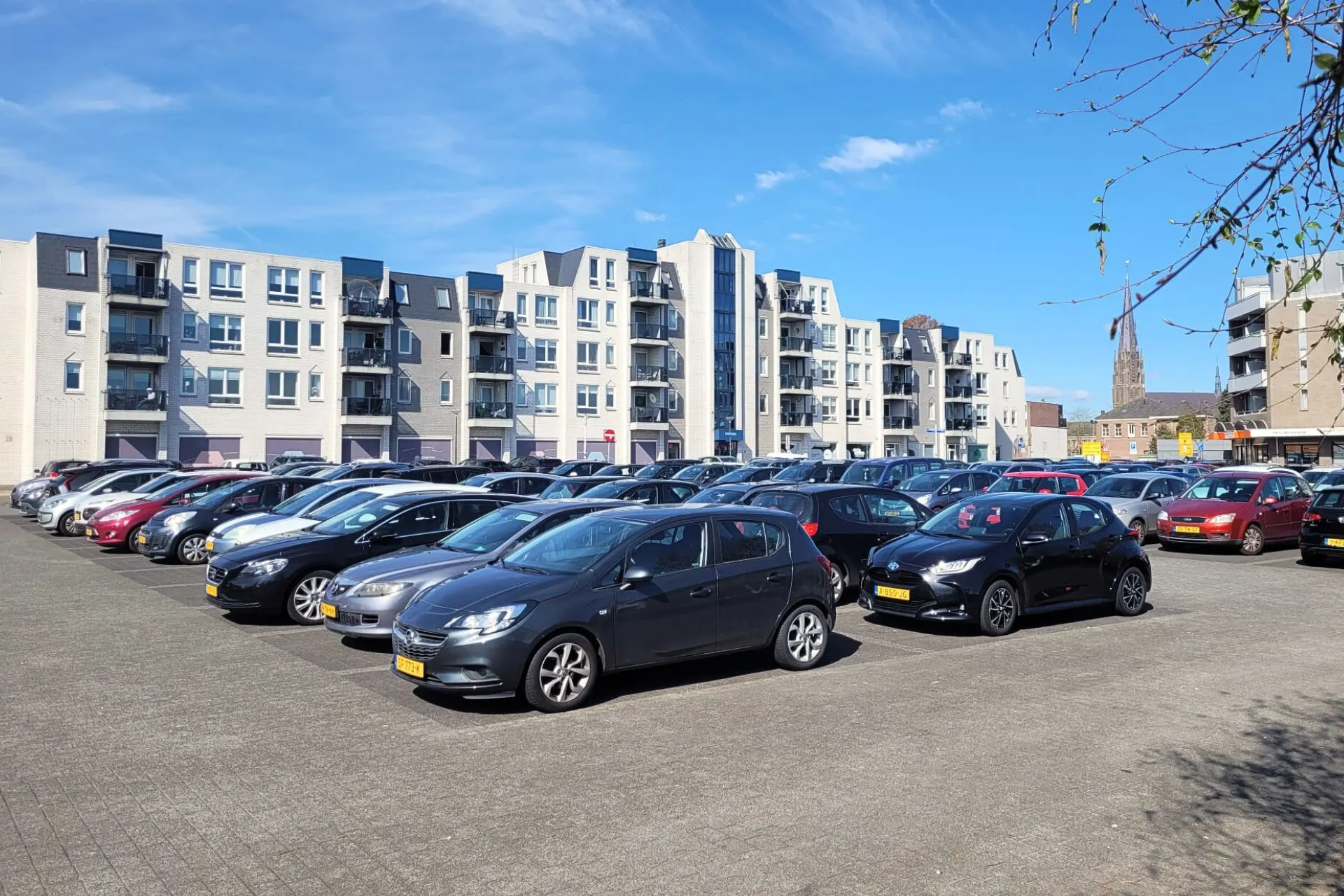 Parkeerplaats stadhuisplein in Veghel op een zonnige dag met blauwe lucht; meerdere rijen met parkeerplaatsen. Dit terrein heeft geen blauwe zone en biedt gratis parkeren zonder tijdslimiet. Vanaf hier is het 2 minuten lopen naar Travelcenter de Wit Veghel.