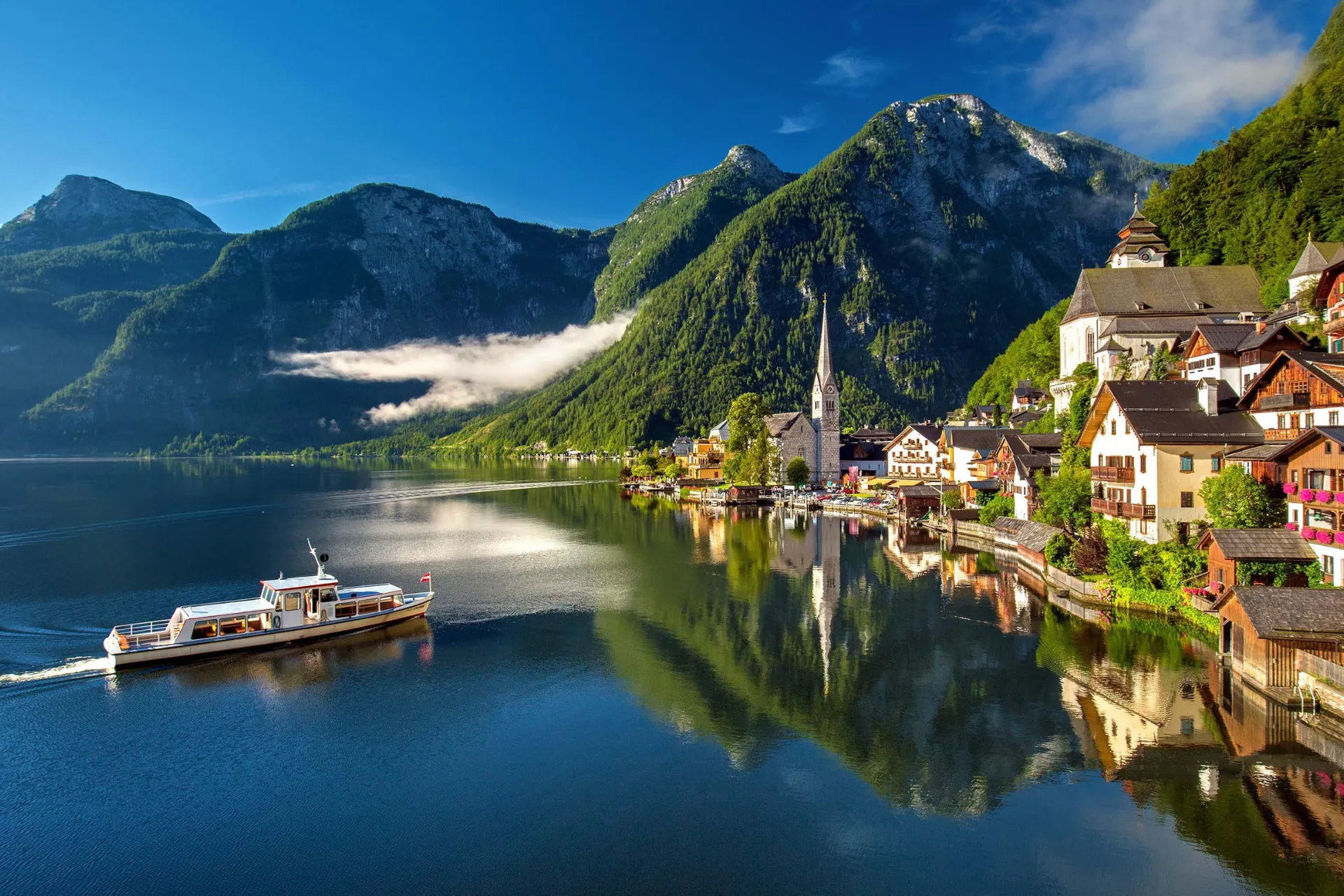 Schilderachtig uitzicht op Hallstatt in Oostenrijk, gelegen aan een helderblauw meer omringd door imposante Alpenbergen en traditionele houten huizen. Een boot vaart over het rustige water, terwijl de historische kerk en kleurrijke gevels weerspiegelen in het meer. Populaire bestemming voor natuur, cultuur en fotografie. Ontdek idyllische landschappen, bergdorpen en UNESCO-werelderfgoed in Oostenrijk.