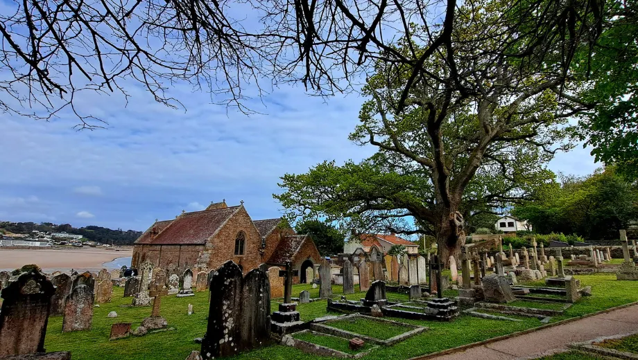 Historische begraafplaats met oude grafstenen en een grote boom die schaduw biedt over het pad en de graven. Op de achtergrond staat een charmant stenen kerkje met puntdak, gelegen nabij een zandstrand en kustlijn. De groene grasvelden en klassieke architectuur creëren een serene en authentieke sfeer, ideaal voor liefhebbers van geschiedenis, cultuur en fotografie. Perfecte locatie om historische monumenten en natuurlijke schoonheid te ontdekken in een rustige omgeving.