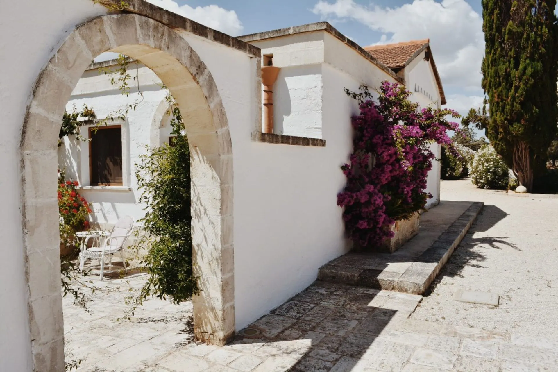 Witte stenen boog en binnenplaats van Masseria Zambardo in Puglia, met bloeiende paarse bougainville en mediterrane begroeiing onder een zonnige hemel.