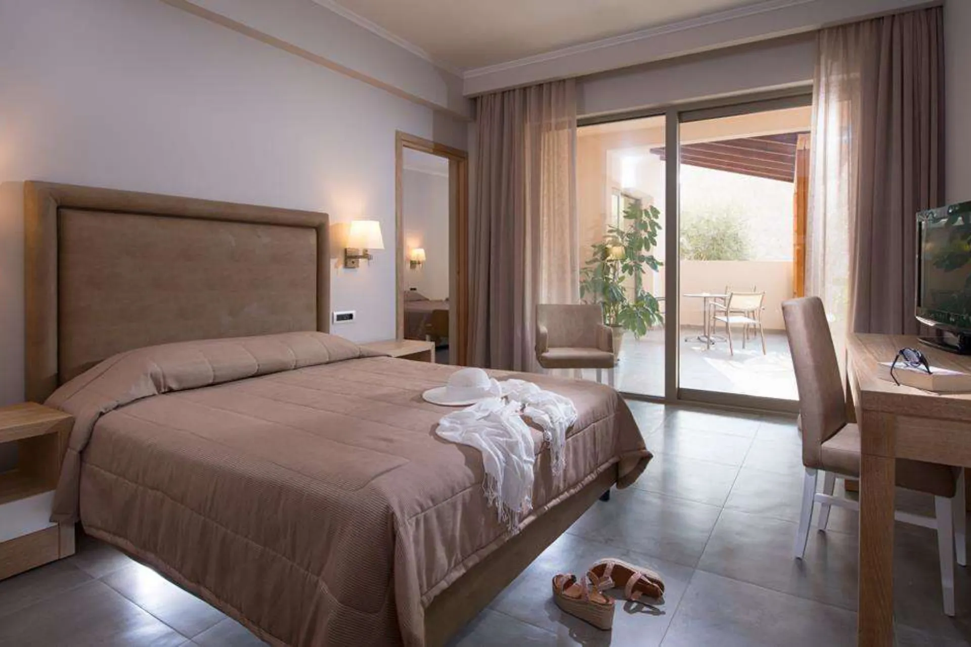 Een stijlvolle hotelkamer met een groot bed bedekt met een beige sprei en witte lakens. Op het bed ligt een opgevouwen badjas, en op de vloer staan sandalen, wat een ontspannen vakantiesfeer oproept. De kamer heeft een neutrale inrichting met houten meubels, een bureau met stoel en een flatscreen-tv. Grote glazen schuifdeuren leiden naar een terras met een tafel en stoelen, omgeven door lichte gordijnen die zacht het zonlicht filteren. De setting straalt comfort en elegantie uit.