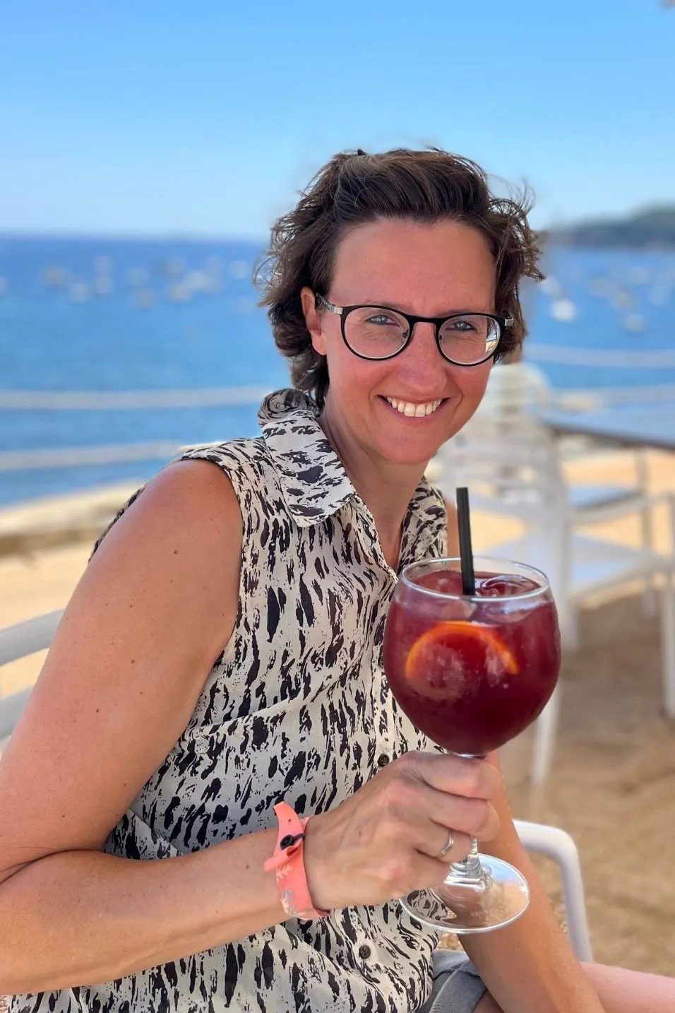 Persoon zit op een terras aan zee onder een parasol en houdt een glas rode sangria vast. Op de achtergrond zijn witte stoelen, tafels en een helderblauwe zee met boeien zichtbaar, samen met een groene kustlijn.
