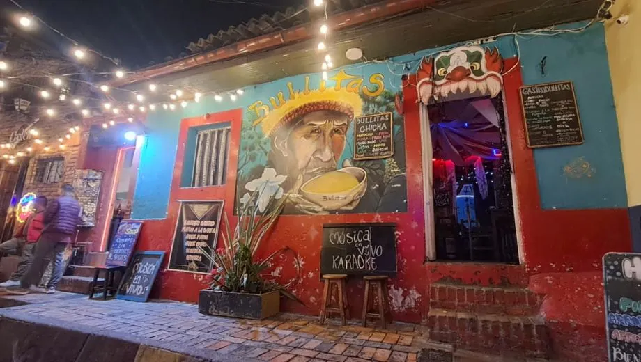 Kleurrijke gevel van een bar met muurschilderingen, lichtslingers en borden met teksten zoals “Música en vivo Karaoke”, gelegen aan een straat met stenen trottoir.