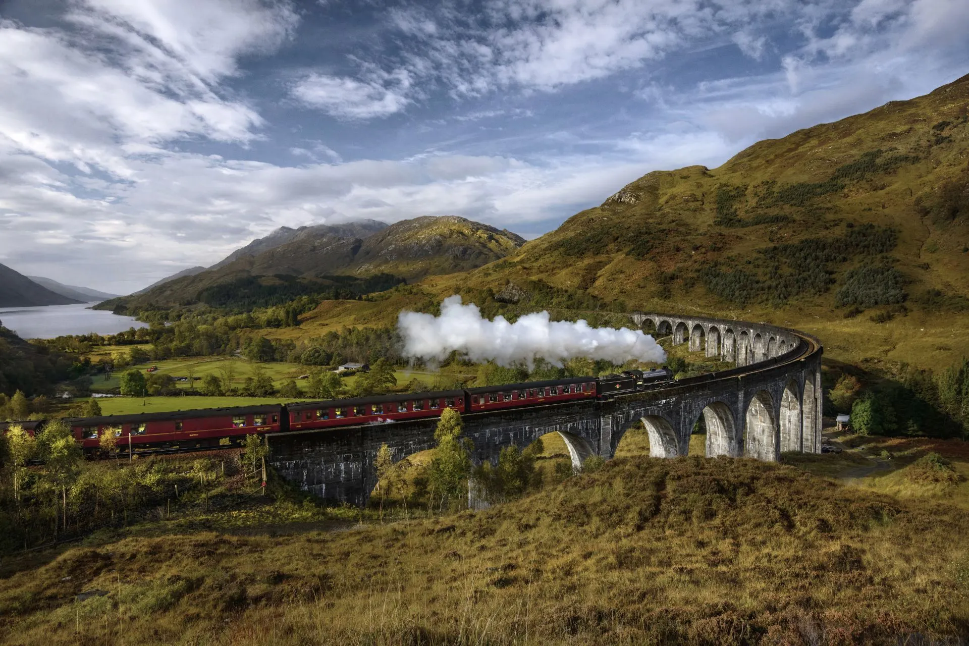 Zwarte stoomtrein met rode wagons rijdt over het iconische Glenfinnan-viaduct te midden van een schilderachtig, groen heuvellandschap.