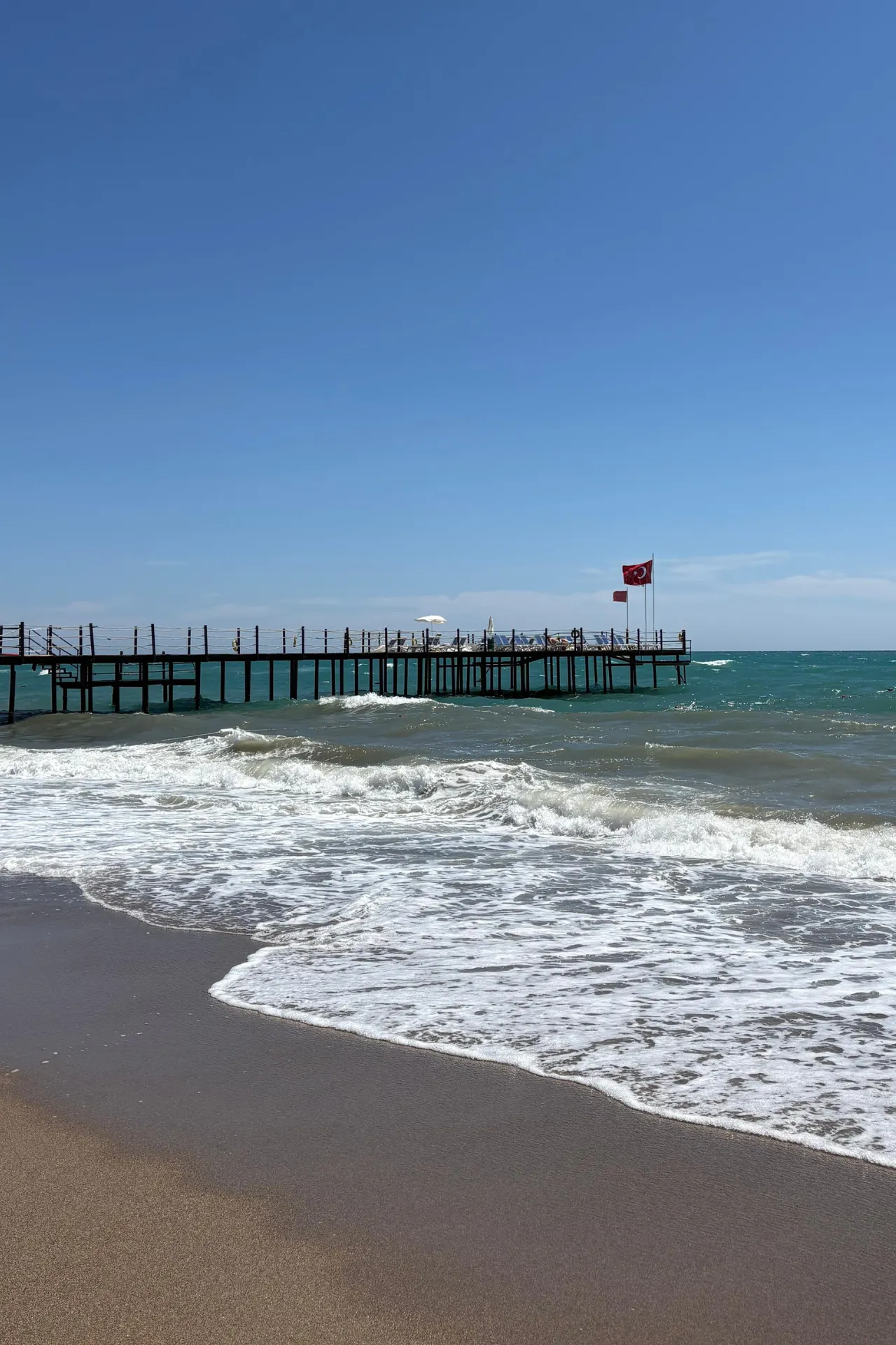 Het privéstrand van Sunmelia Beach Resort aan de Turkse Rivièra met zacht zand, golven van de Middellandse Zee en een lange pier die de zee in loopt. Gasten kunnen hier genieten van zon, strand en uitzicht over de kust van Turkije tijdens een ontspannen all inclusive vakantie nabij Side.
