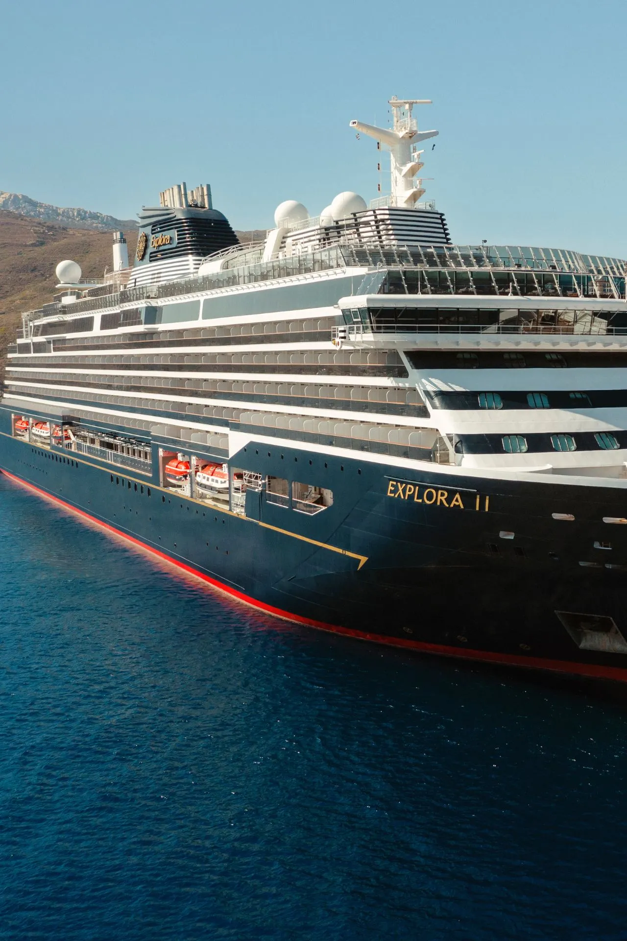 Het moderne cruiseschip Explora II van Explora Journeys ligt aangemeerd in een helderblauwe zee, omringd door een bergachtig landschap. Het schip straalt elegantie en comfort uit met meerdere dekken, ruime balkons en luxe voorzieningen. Deze Luxe cruise biedt een unieke ervaring waarbij stijl en verfijning centraal staan, ideaal voor reizigers die op zoek zijn naar ontspanning en avontuur. Het design en de details benadrukken de premium kwaliteit van deze reis op zee.