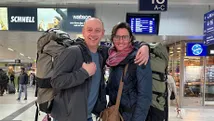 Onze Reisspecialist Susan en haar. Een lachend stel met grote backpacks op een treinstation, klaar voor een avontuurlijke reis naar München.